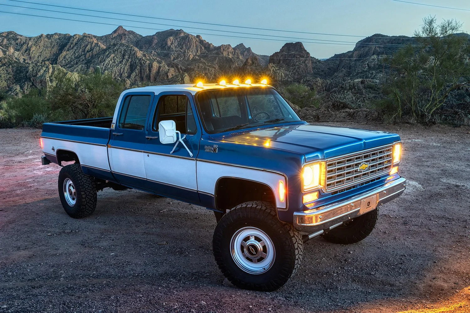 A blue and white Chevrolet pickup truck with its headlights and roof lights on, parked on a dirt surface in a desert with mountains and sparse vegetation in the background.