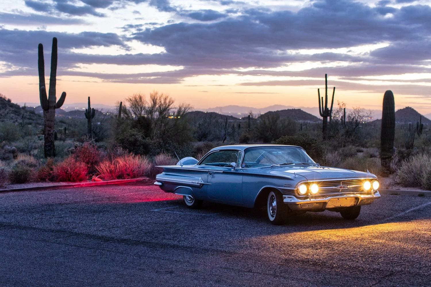 A vintage blue Chevrolet car parked in a desert landscape at sunset, with cacti and desert shrubs in the background.