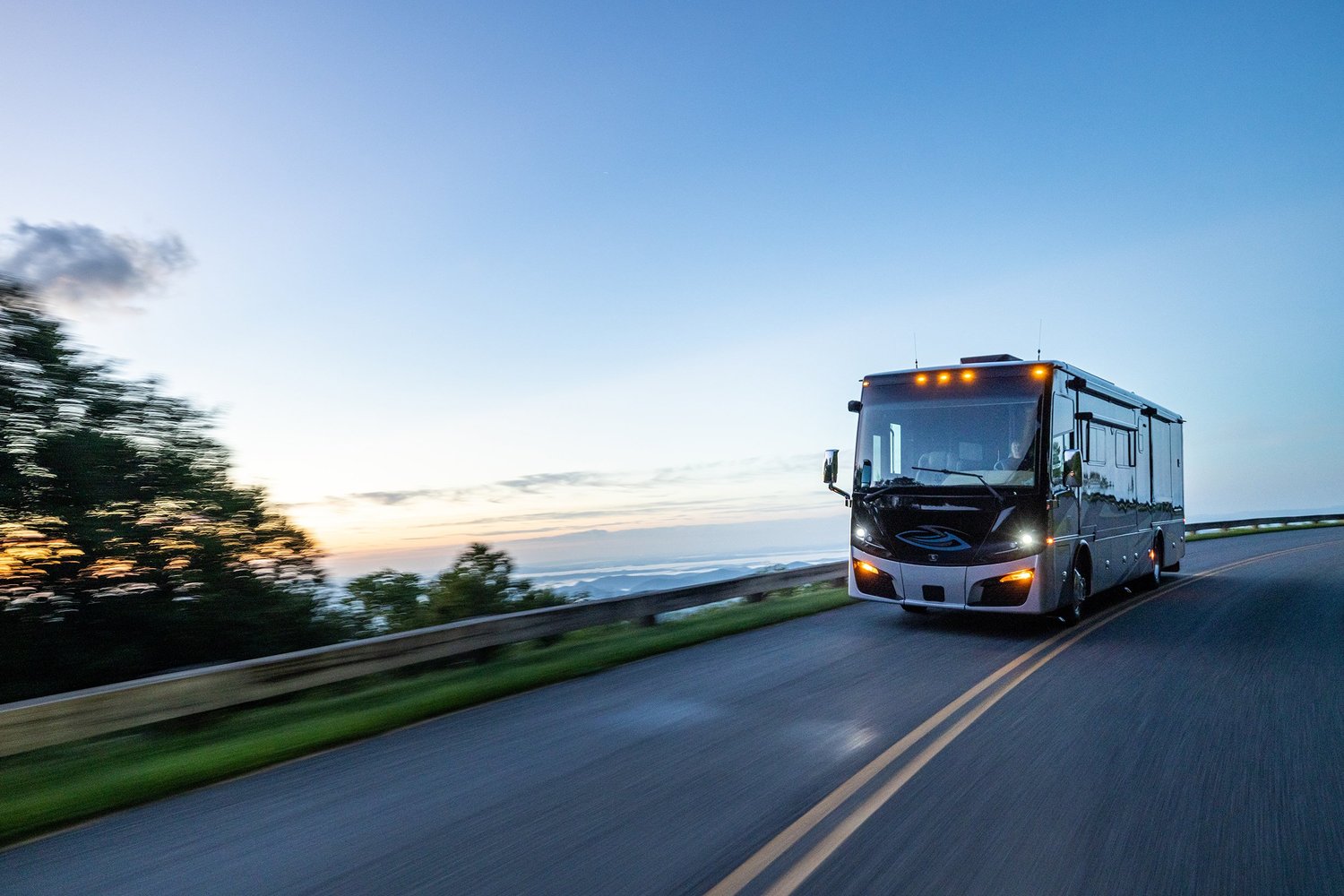 A modern bus traveling on a winding road at sunset with trees and a scenic view in the background.