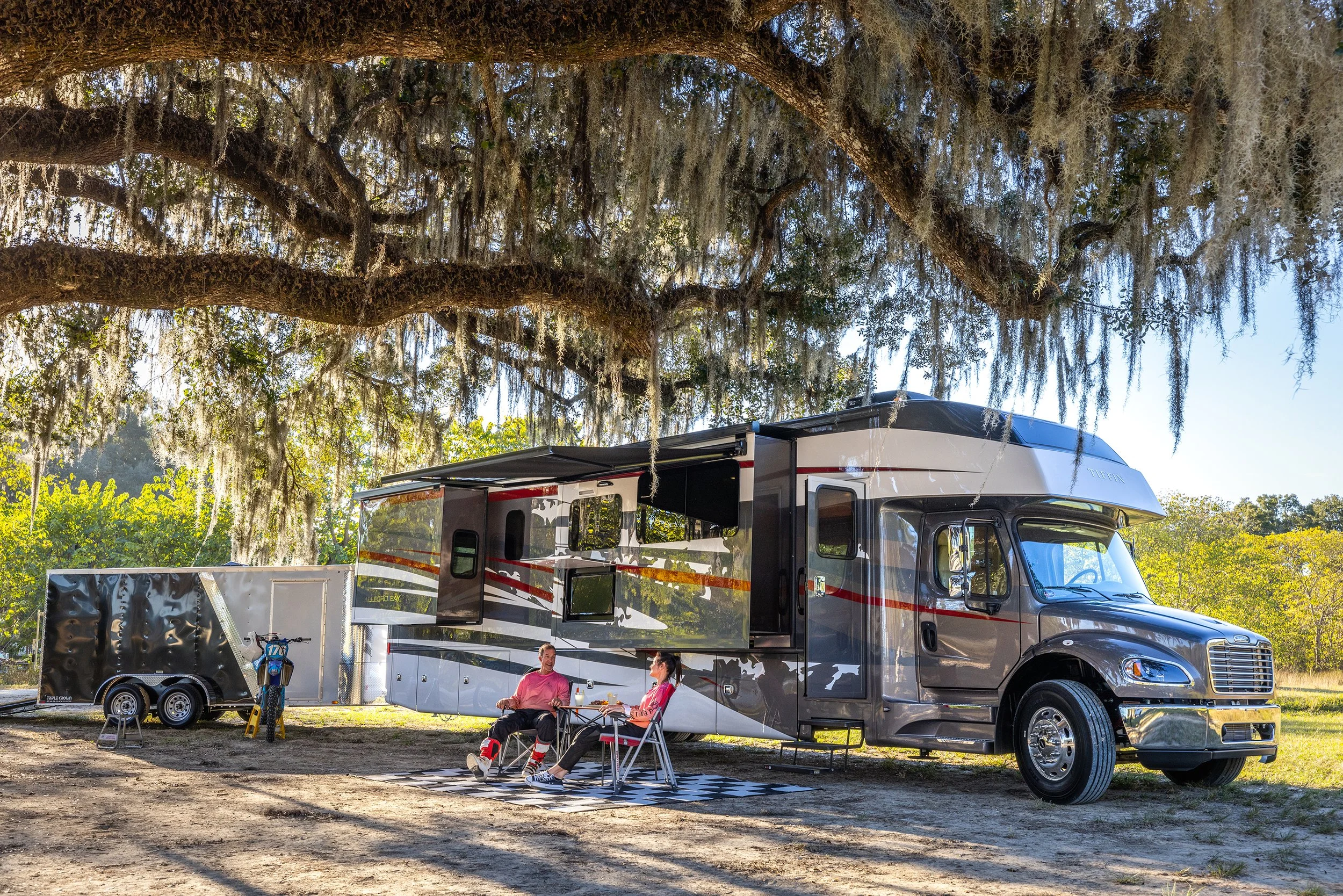 A large motorhome parked under a sprawling tree with Spanish moss, with two people sitting outside at a small table, and a trailer attached to the motorhome.