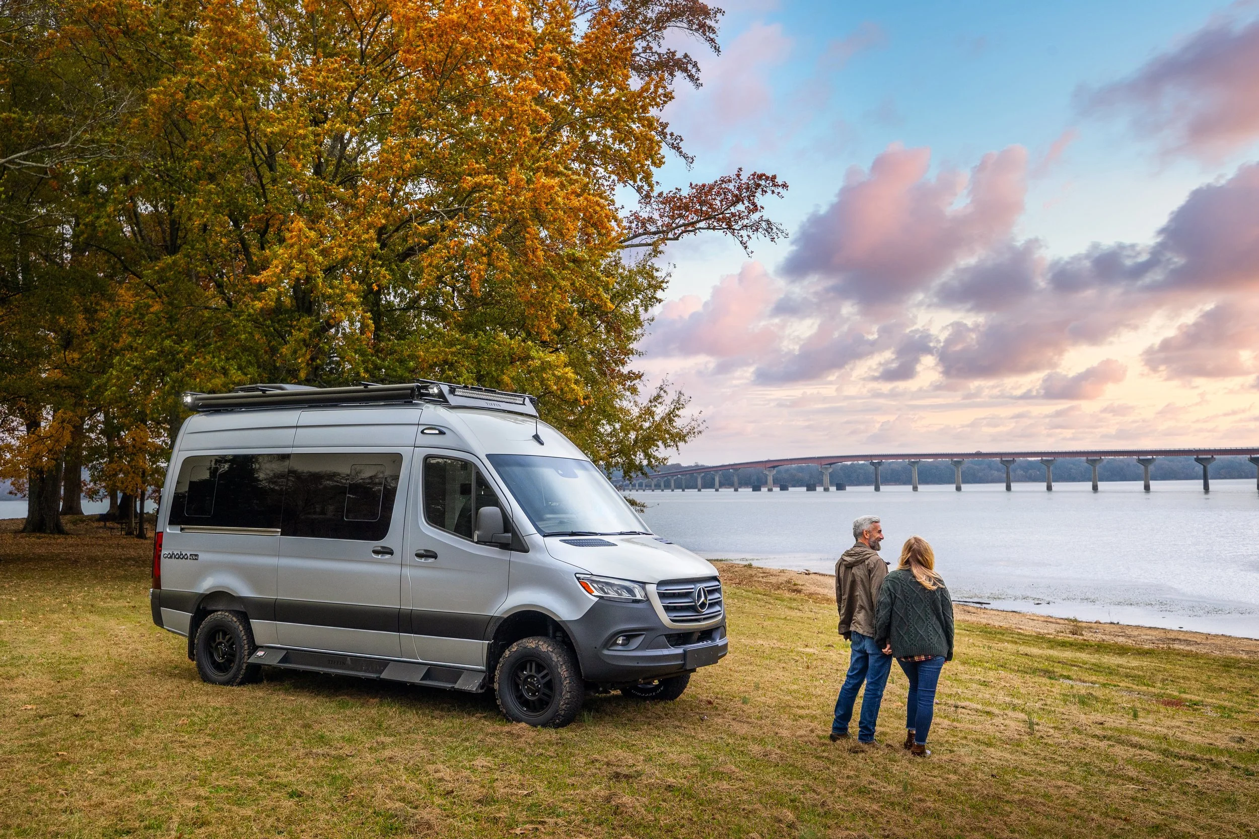 A gray camper van parked on a grassy area near a body of water, with two people walking hand in hand along the shoreline. There are trees with fall foliage and a long bridge across the water in the background, under a cloudy evening sky.