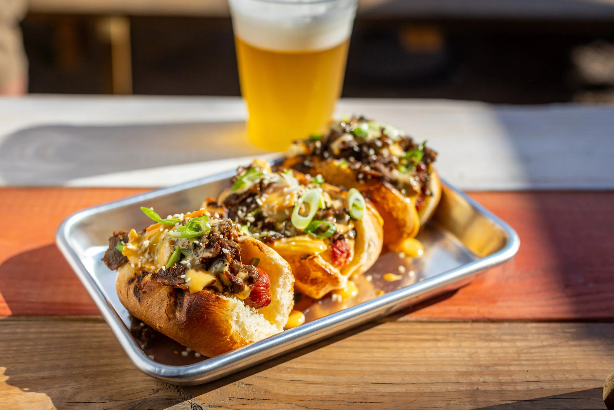 Three tacos with beef, cheese, green onions, and sauce served on a metal tray, with a glass of beer in the background on a wooden table.