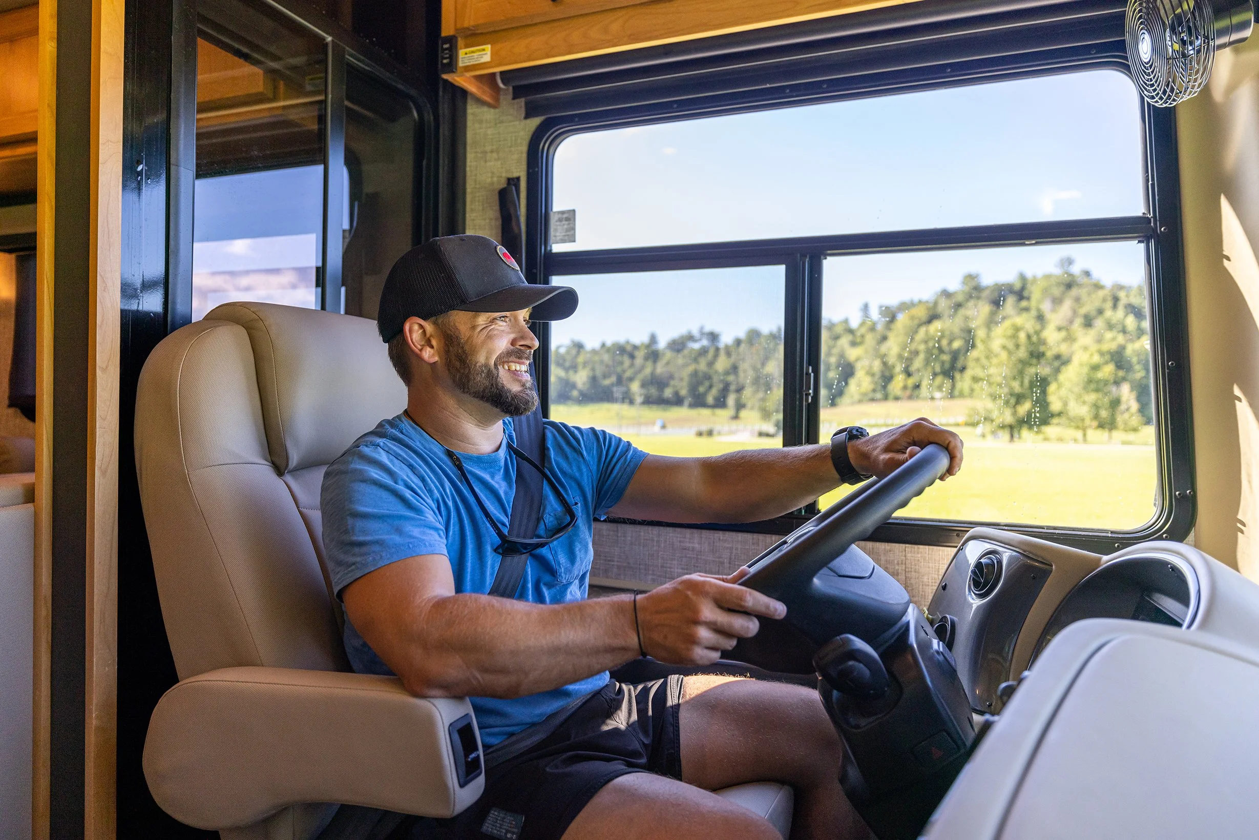A man with a beard and black cap driving a bus, smiling through the window with a scenic view of trees and grass.