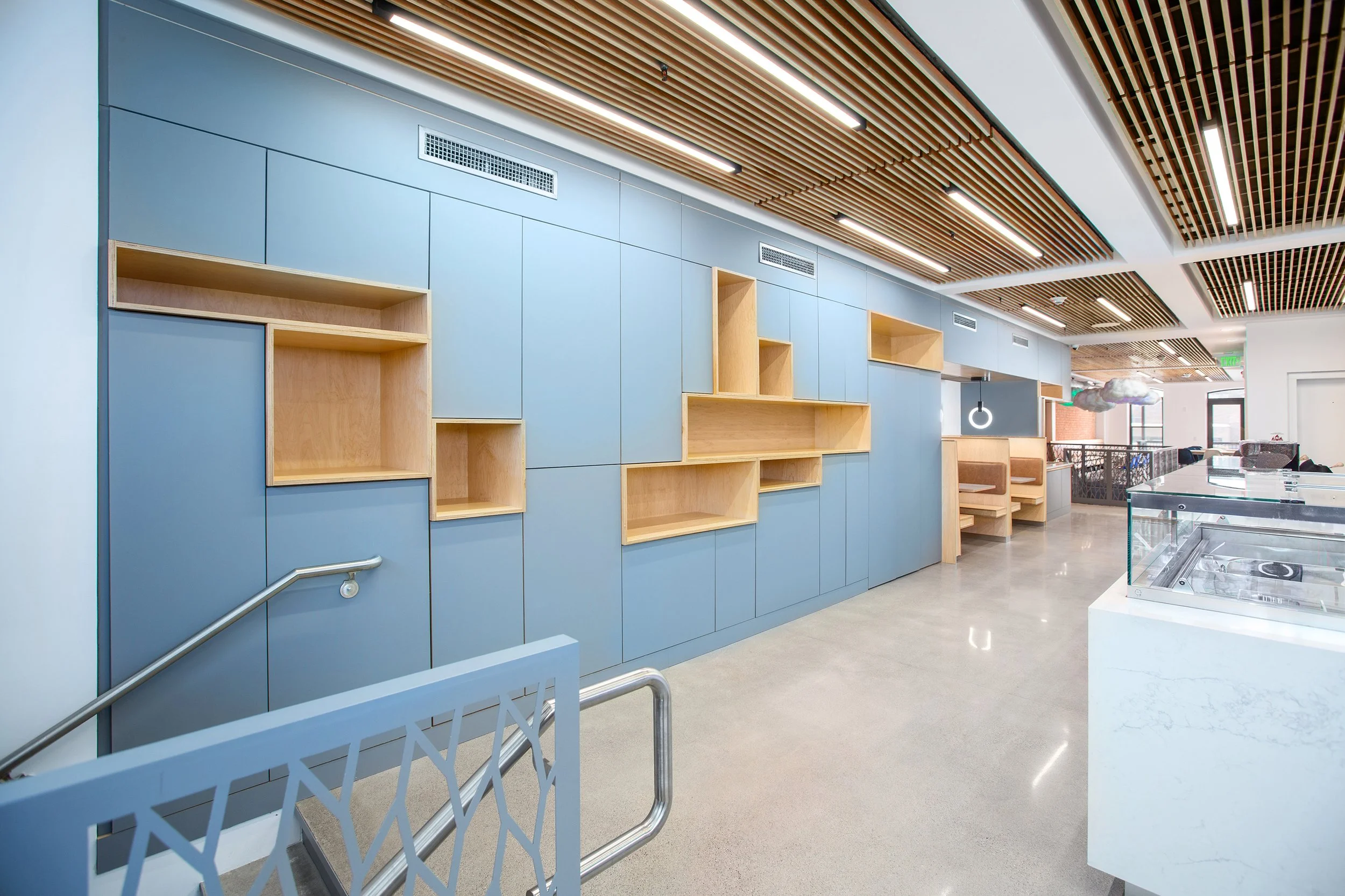 Modern interior space with blue wall panels, wooden shelves, a ceiling with wooden slats, and seating areas in the background.