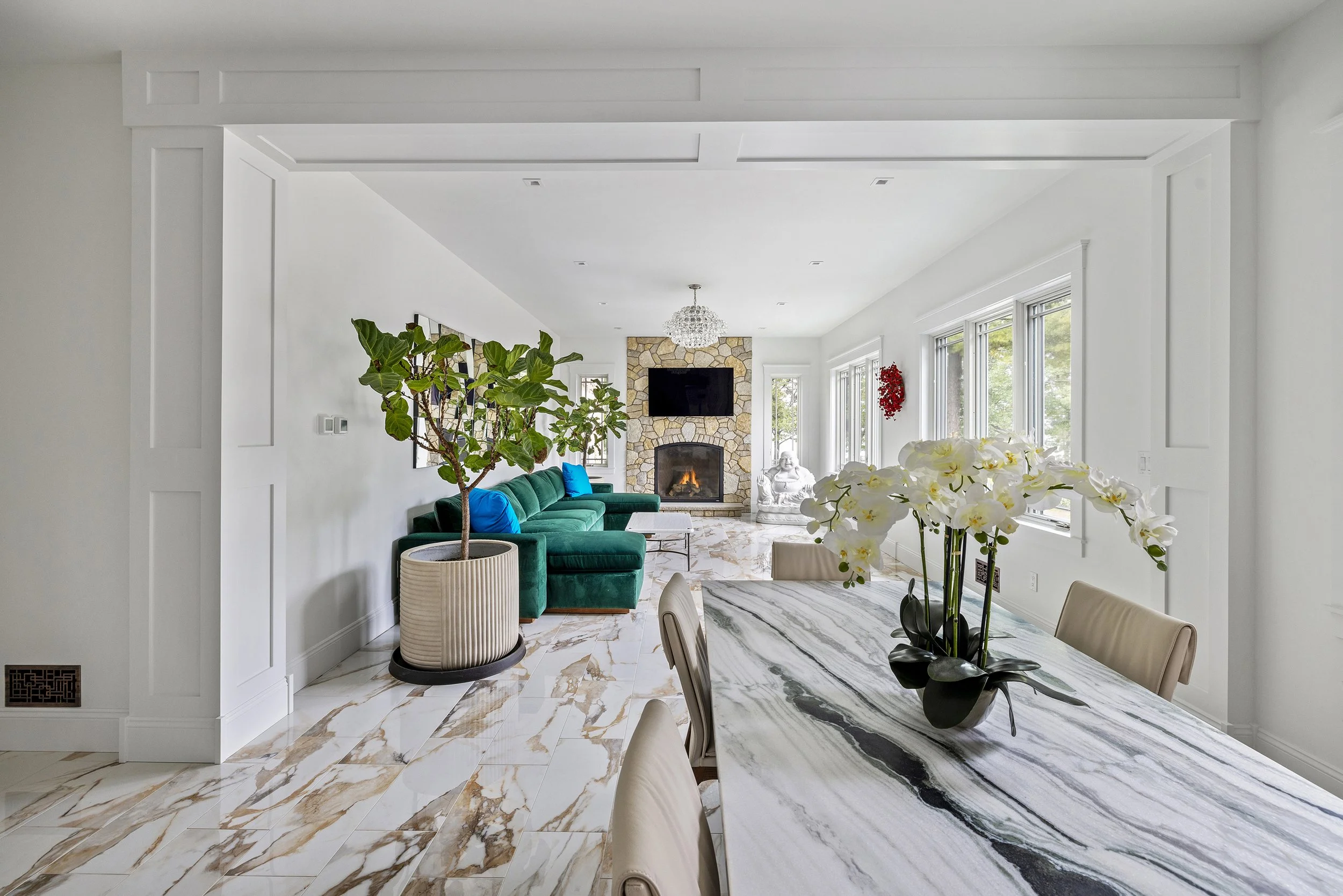 Modern living room with white walls, marble floors, green velvet sofa, large potted plant, fireplace with stone surround, mounted TV, and a chandelier. A dining table with a floral centerpiece is visible in the foreground.