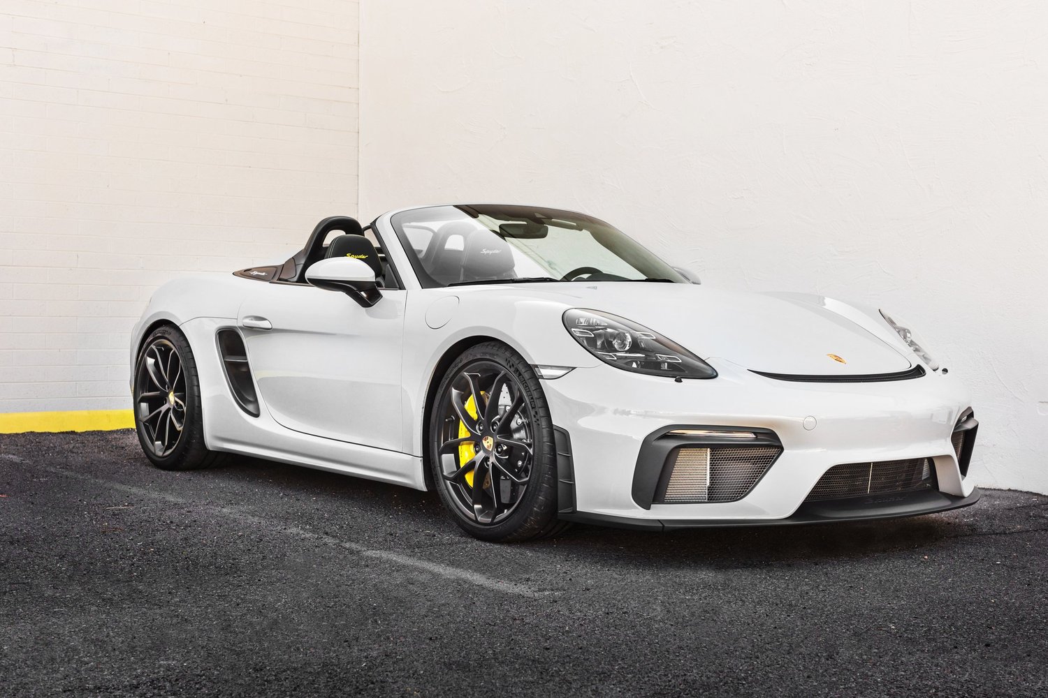 A white Porsche 718 Spyder convertible parked against a plain white wall.