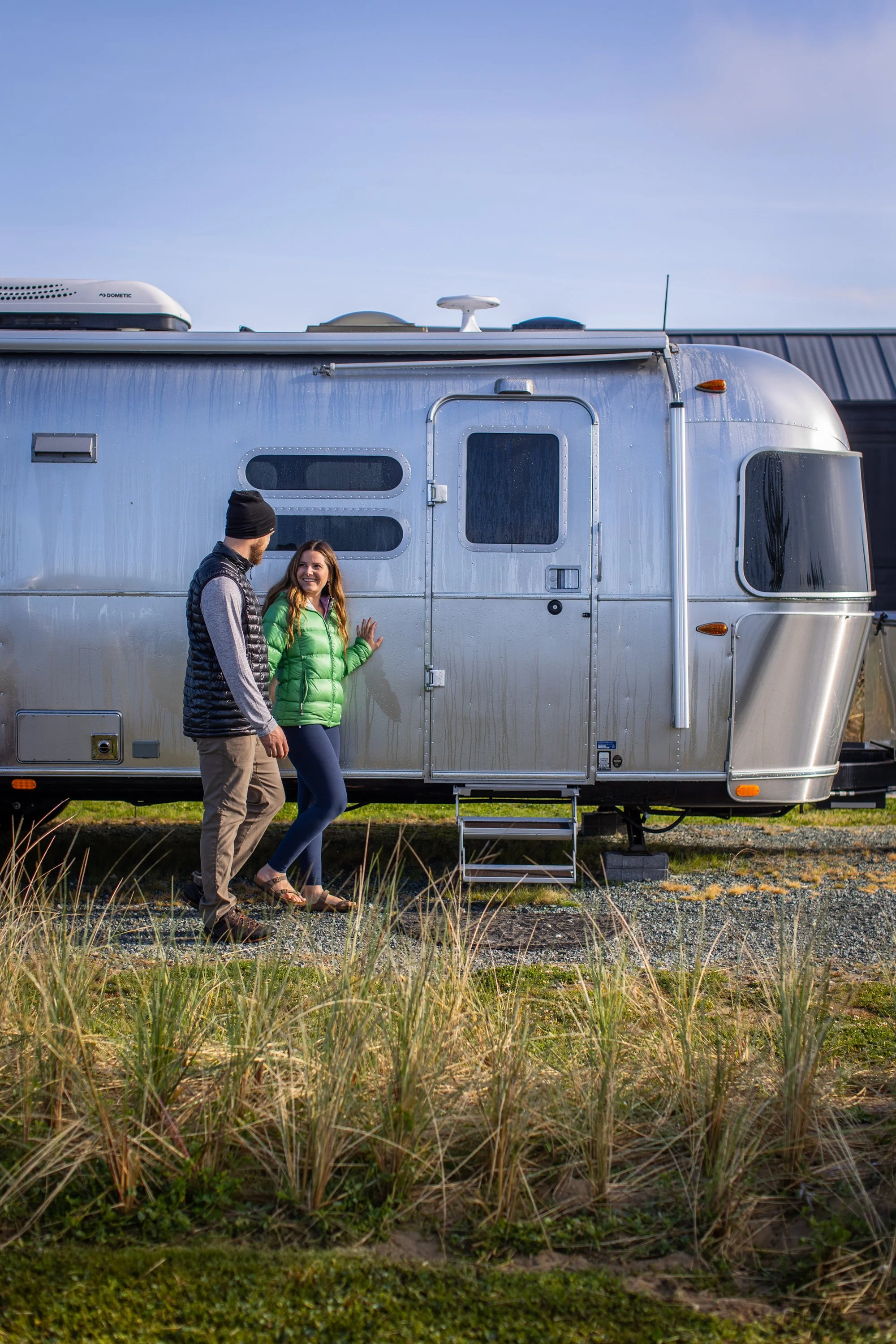 A man and woman standing outside of a silver travel trailer, smiling and talking, with the woman touching the side of the trailer.