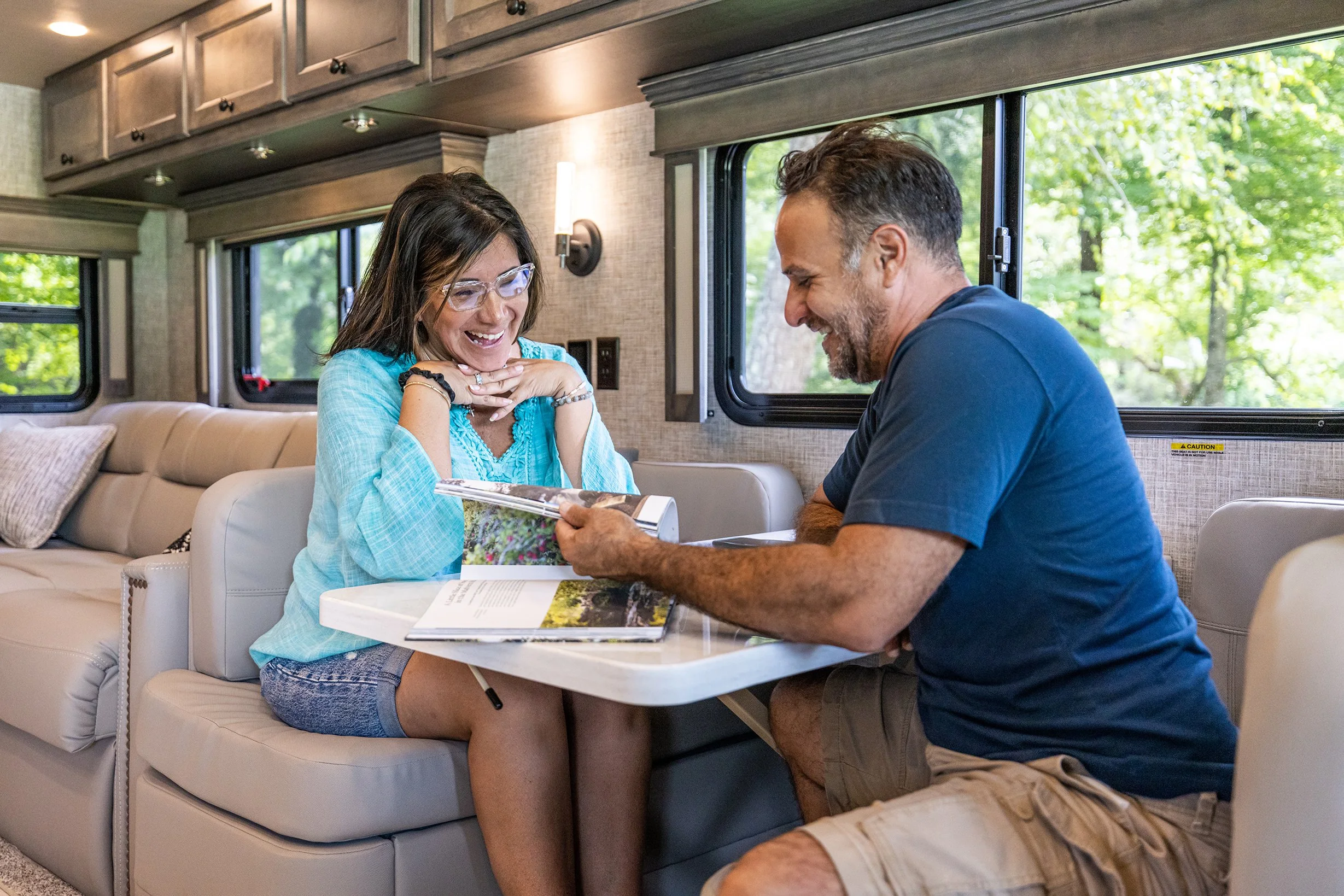 A man and woman sitting at a small table inside an RV, looking through a photo album and smiling, with windows showing green trees outside.