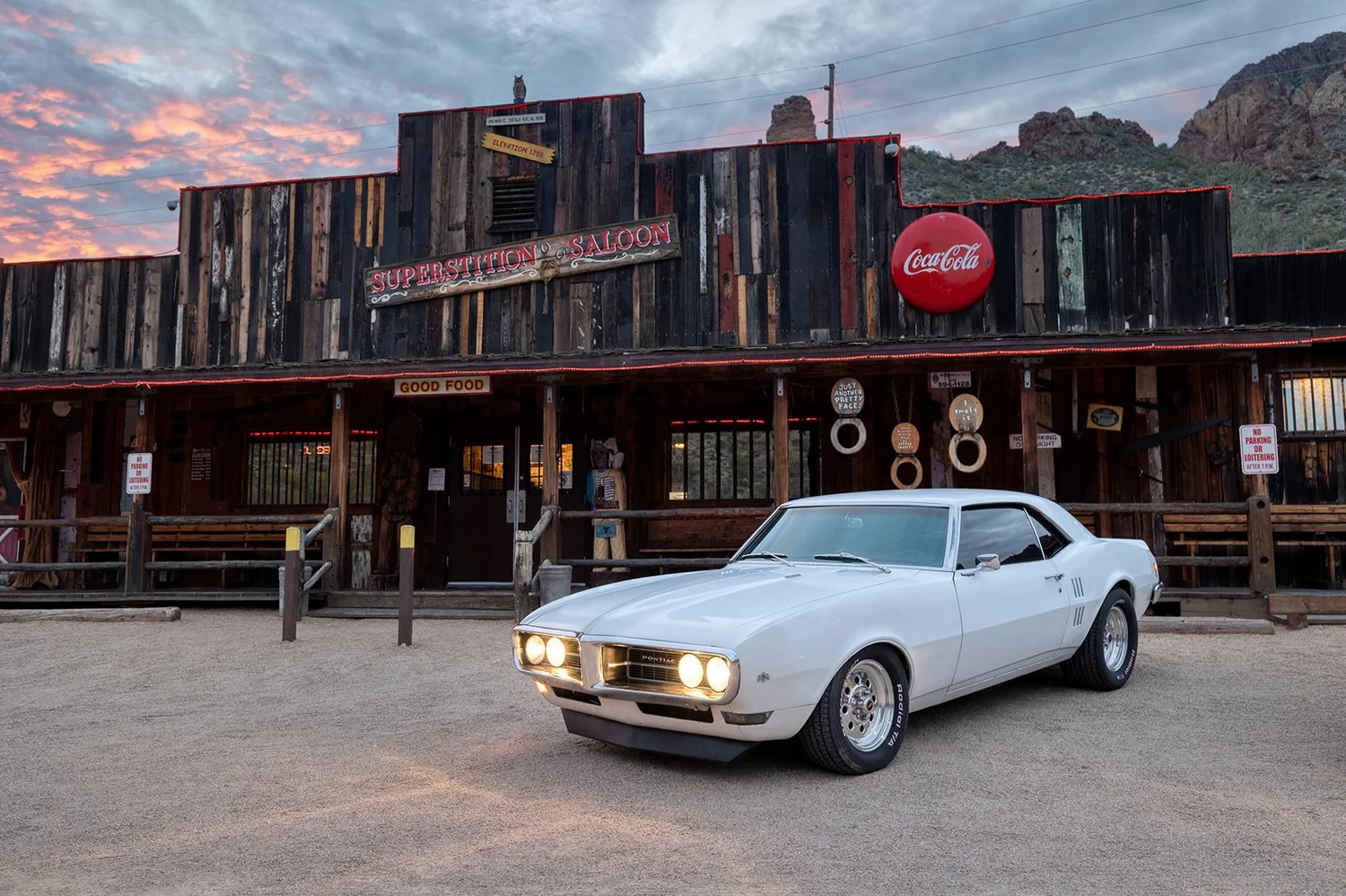 A white classic Pontiac muscle car parked in front of a rustic wooden saloon building with a sign that reads 'Superstition Saloon', at sunset.