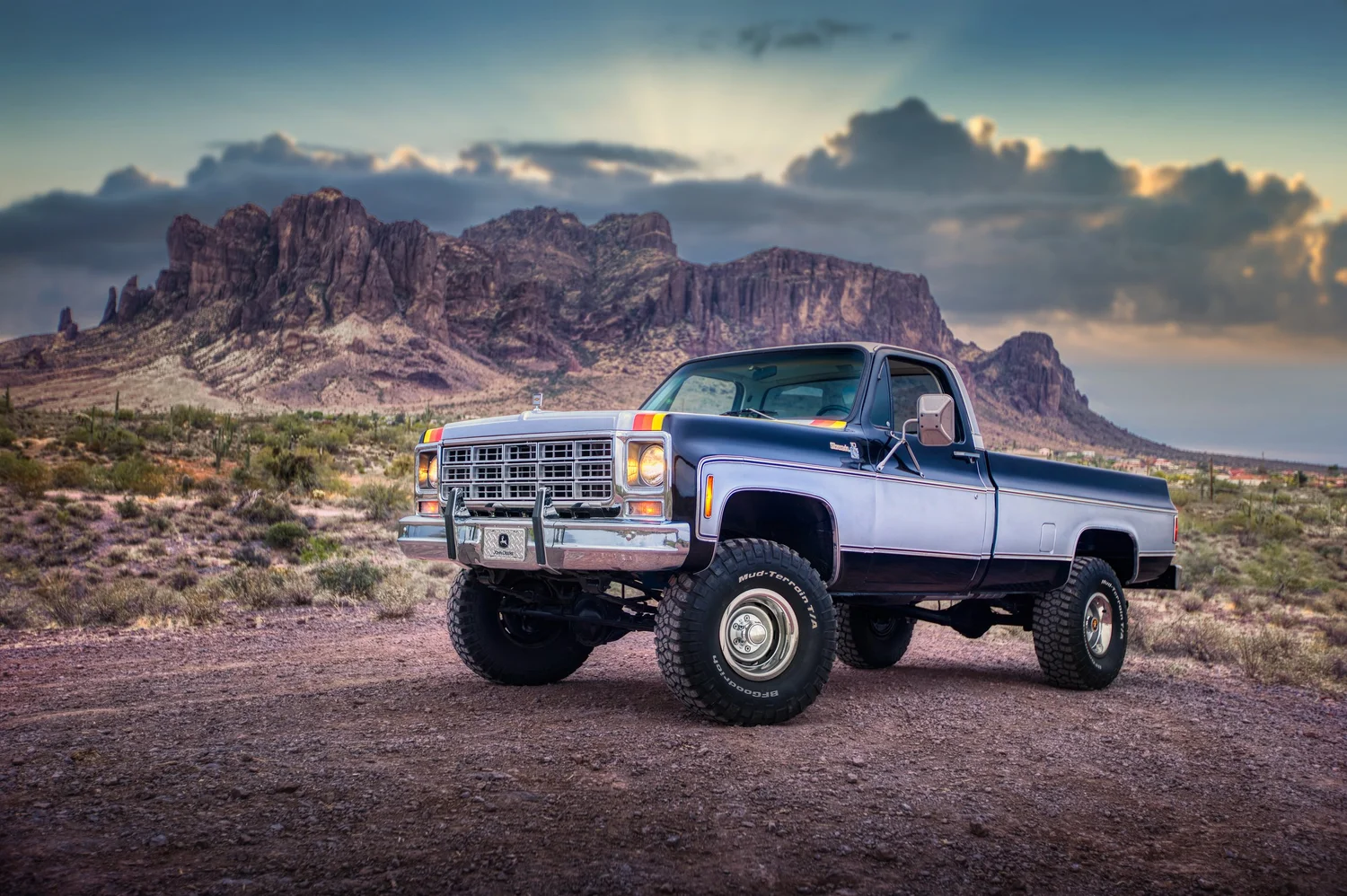 A vintage black and silver pickup truck with large off-road tires parked on a dirt road in a desert landscape with mountains and cloudy sky in the background.
