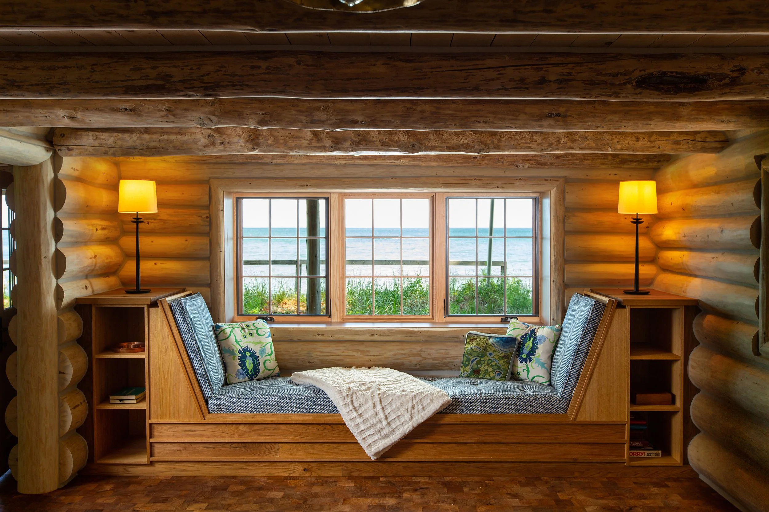 Cozy interior featuring a built-in wooden window seat with patterned pillows, flanked by two lamps, overlooking a body of water through three-pane windows, with log walls and ceiling.