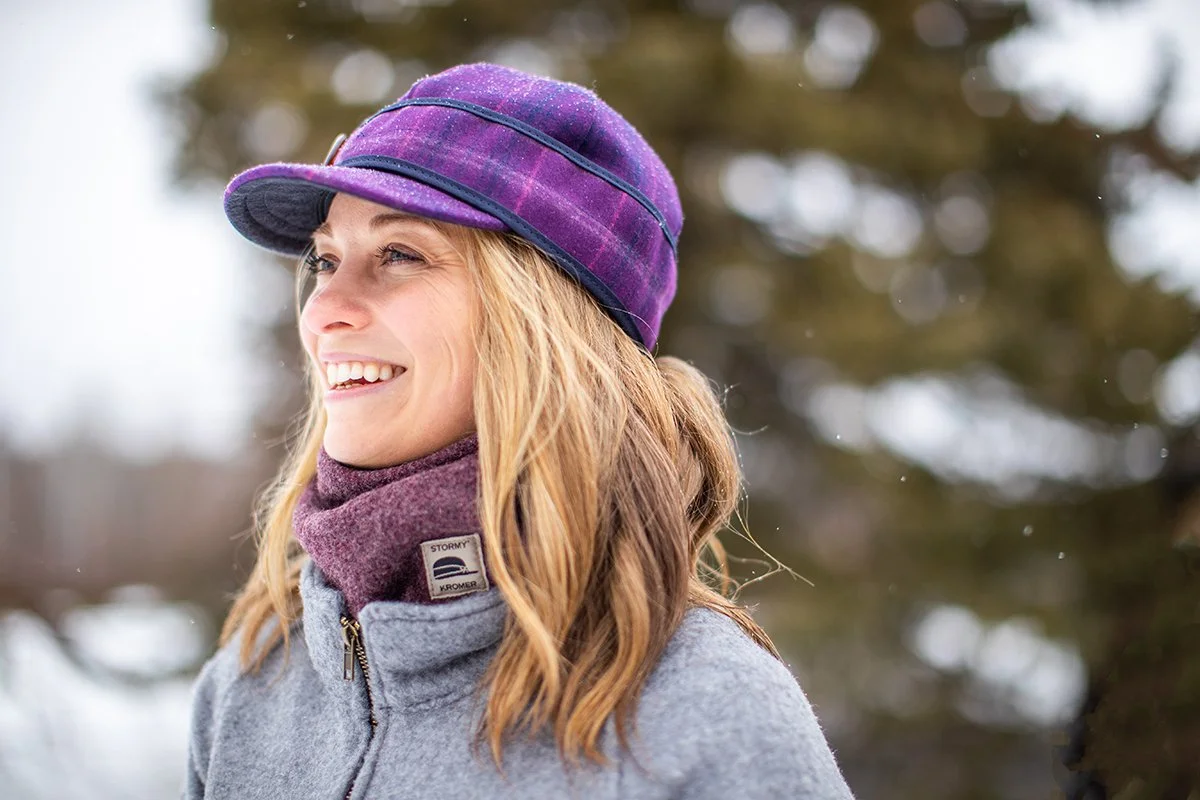 A woman smiling outdoors in winter weather, wearing a purple hat, a gray jacket, and a maroon neck warmer, with snow and trees in the background.