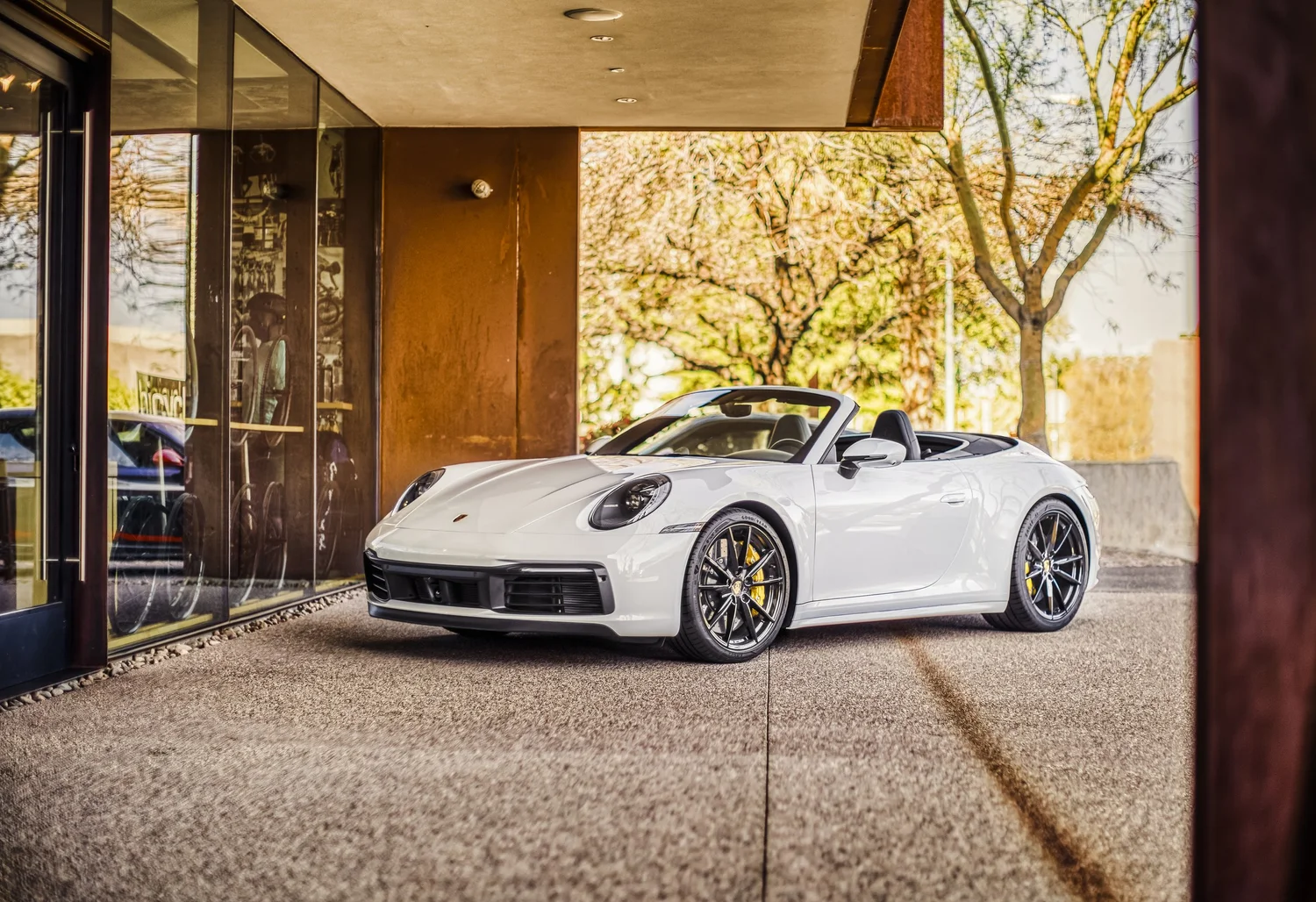 A white Porsche convertible parked outside a building with glass windows, with trees and blue sky in the background.