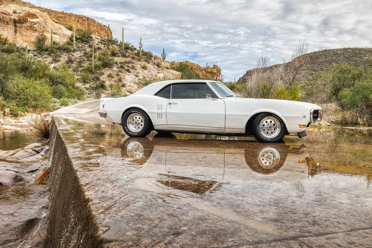 A vintage white car parked on a concrete ledge over a small body of water in a desert landscape with hills, cacti, and sparse vegetation.