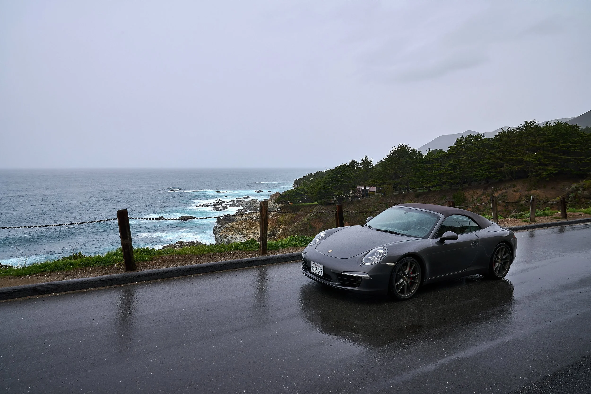 A gray Porsche convertible parked on a wet road near the coast, with an ocean, rocky shoreline, and green trees in the background under an overcast sky.