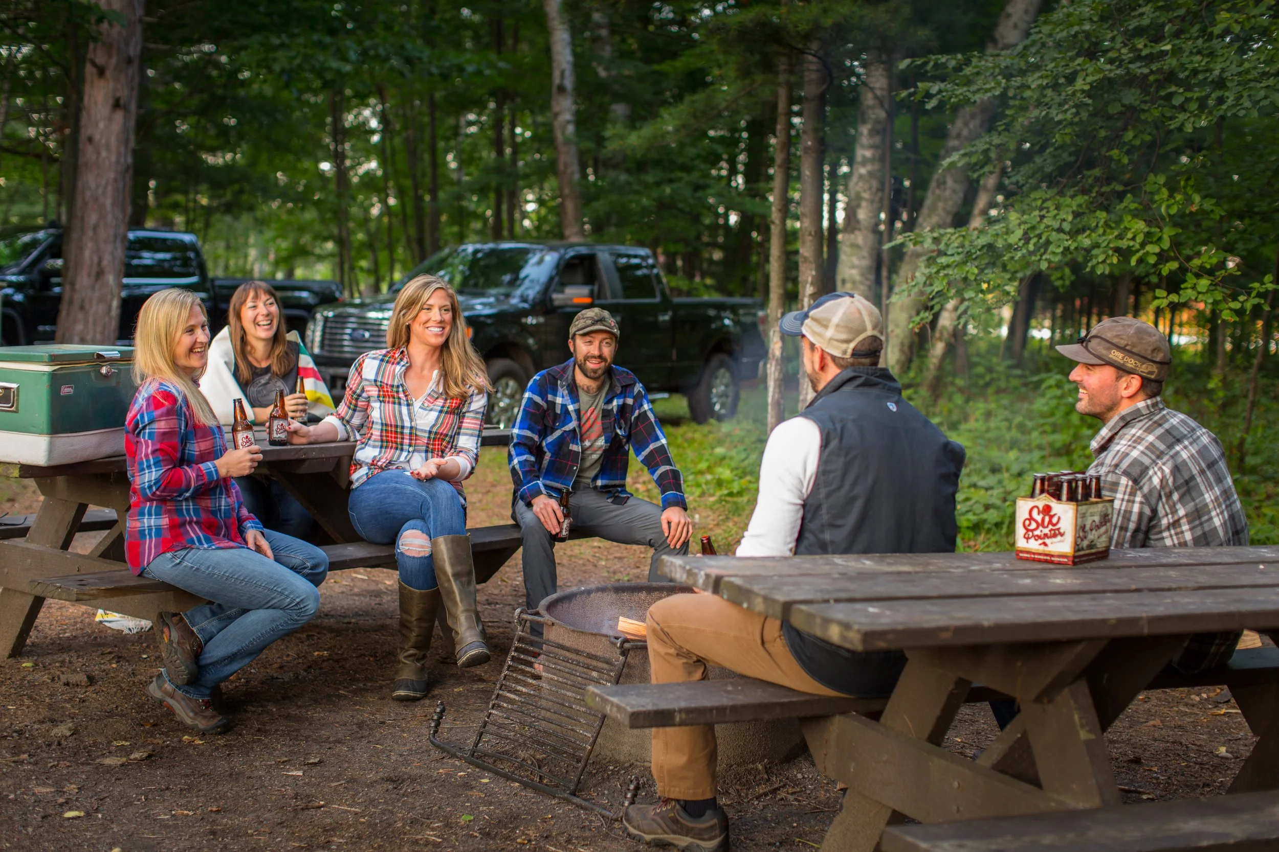 Group of friends sitting around a campfire, laughing and enjoying drinks in a wooded area with trees and parked trucks in the background.