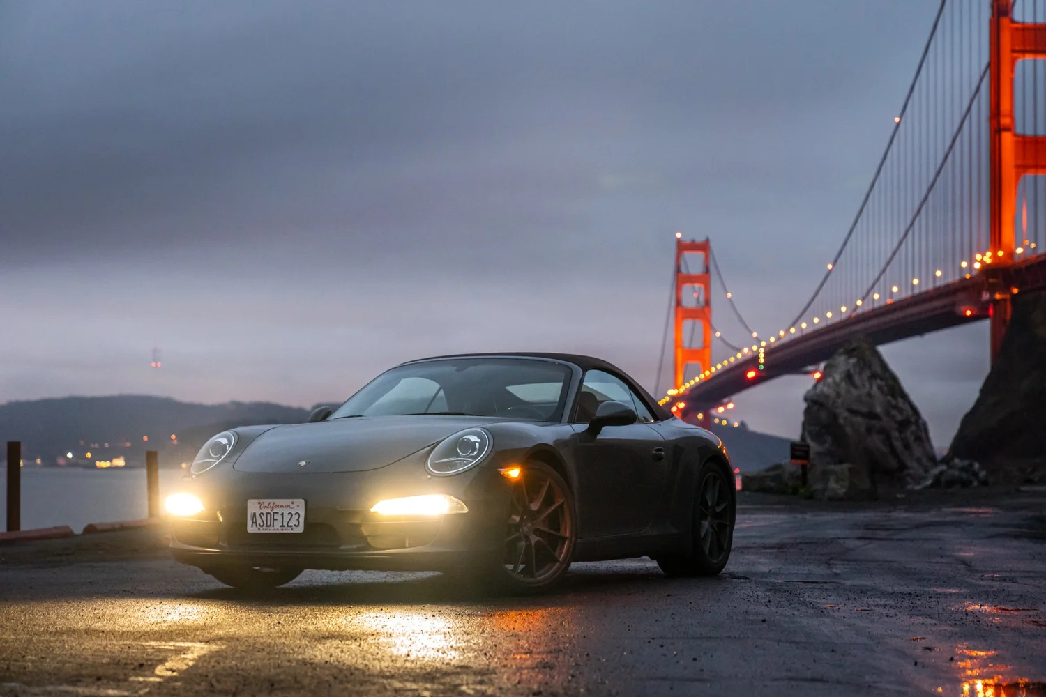 A black Porsche sports car with its headlights on parked near the Golden Gate Bridge at dusk, with a cloudy sky overhead.