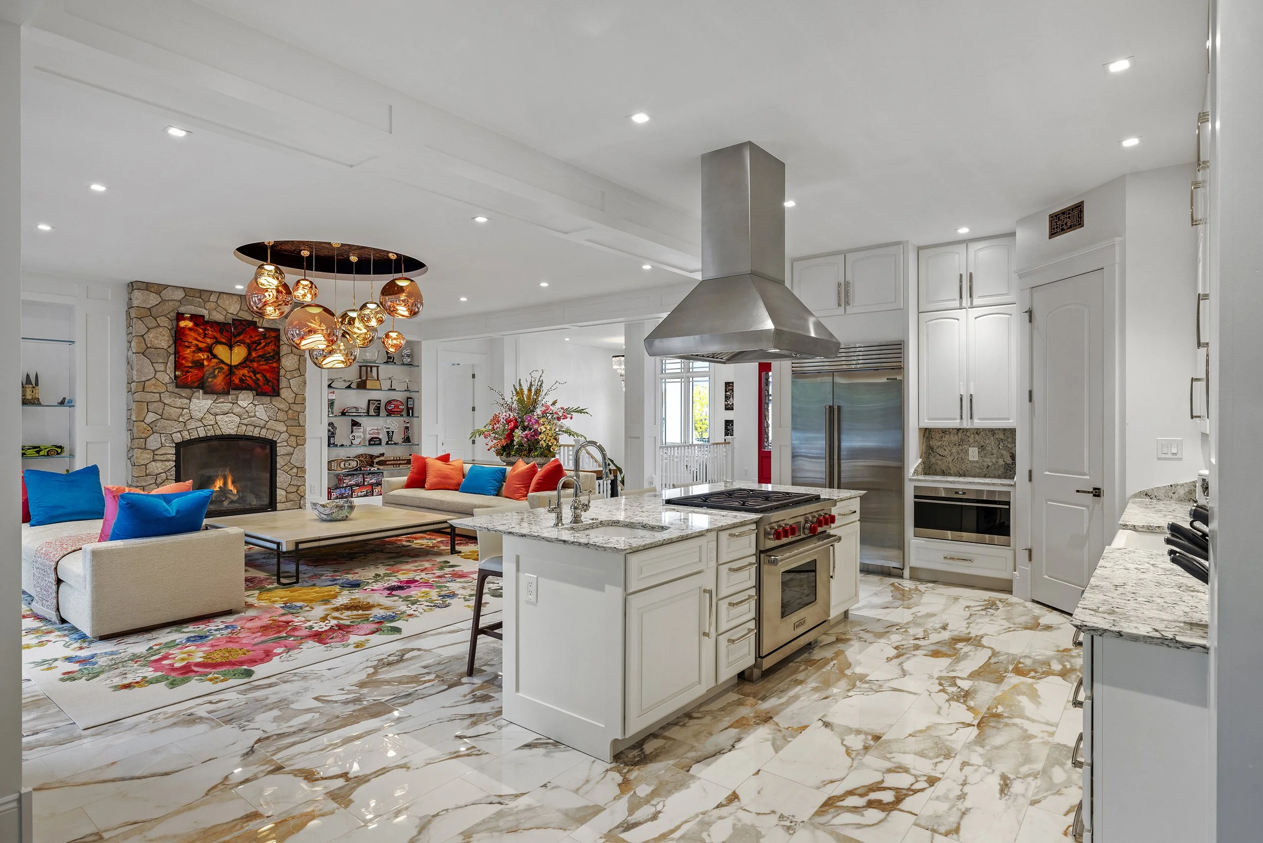 Open-concept kitchen and living room with white cabinets, marble floors, stainless steel appliances, fireplace, colorful cushions, and floral rug.