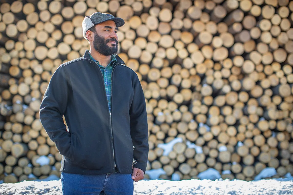 A bearded man wearing a cap, jacket, and plaid shirt standing outdoors in front of a large stack of cut logs.
