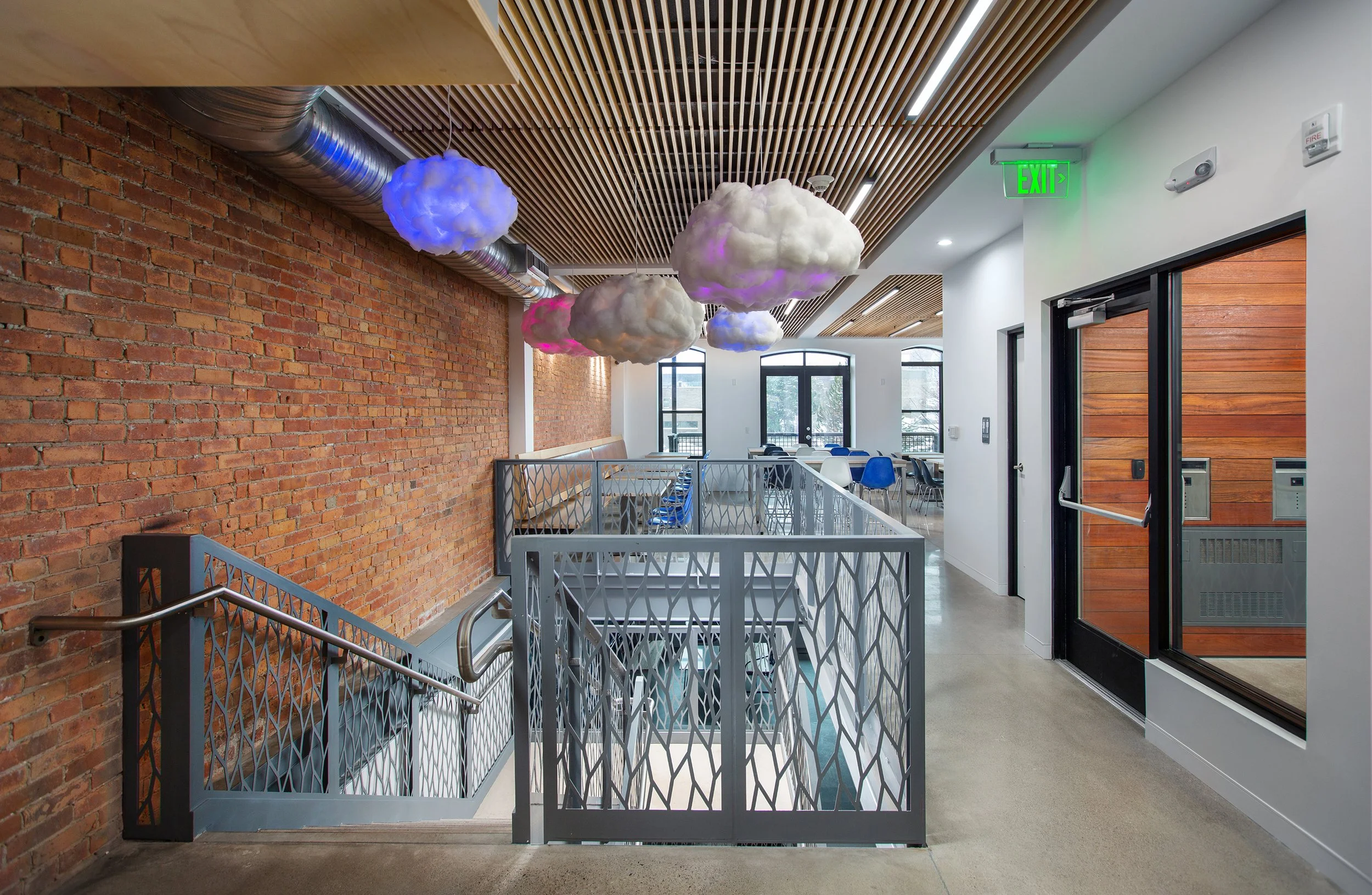 Interior of a modern building with cloud-shaped light fixtures hanging from a wooden ceiling, red brick wall, staircase with metal railing, and a sitting area with blue chairs behind a white railing.