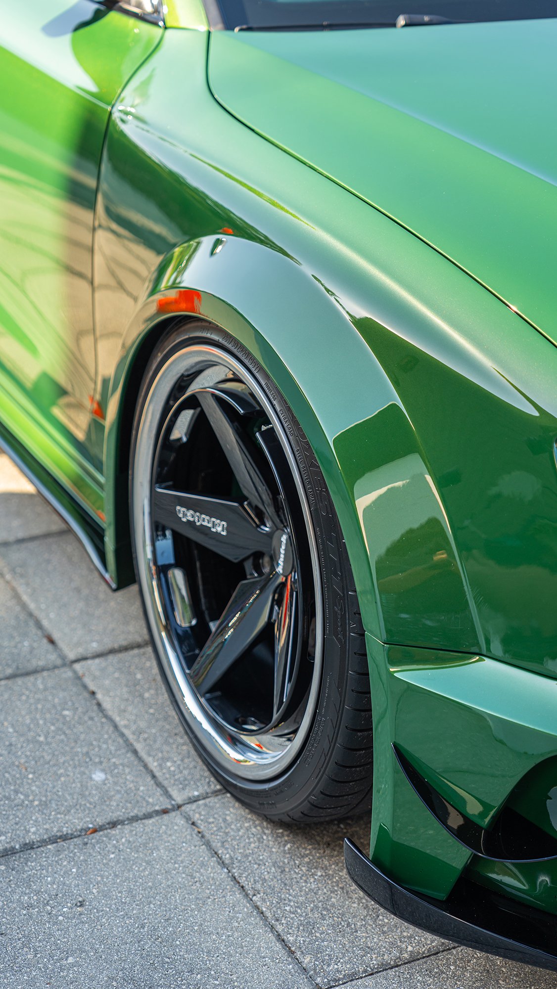 Close-up of a green sports car with black alloy wheel and low-profile tire, parked on a paved surface.