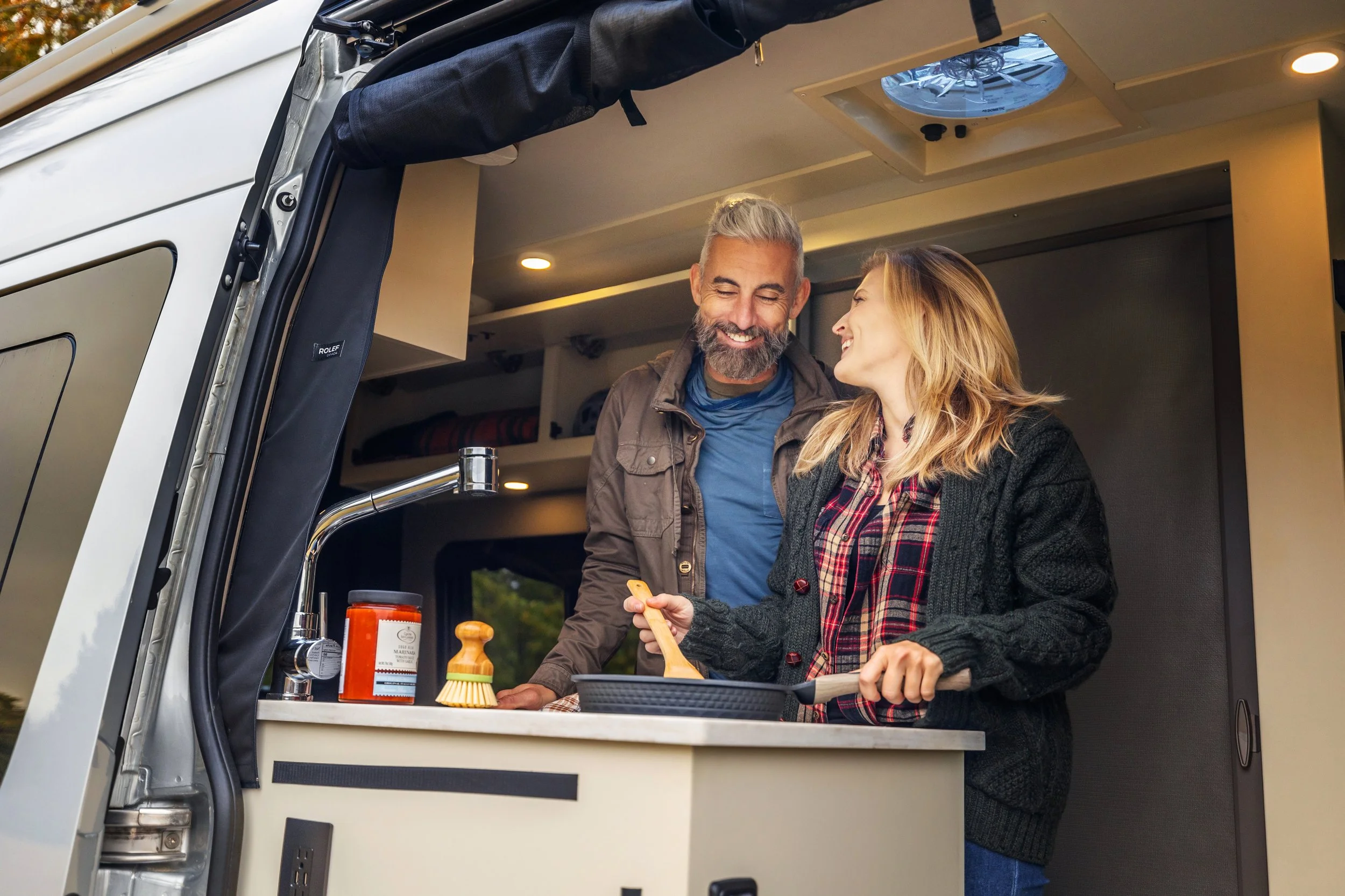 A man and woman cooking and smiling inside a tiny home or camper RV kitchen.