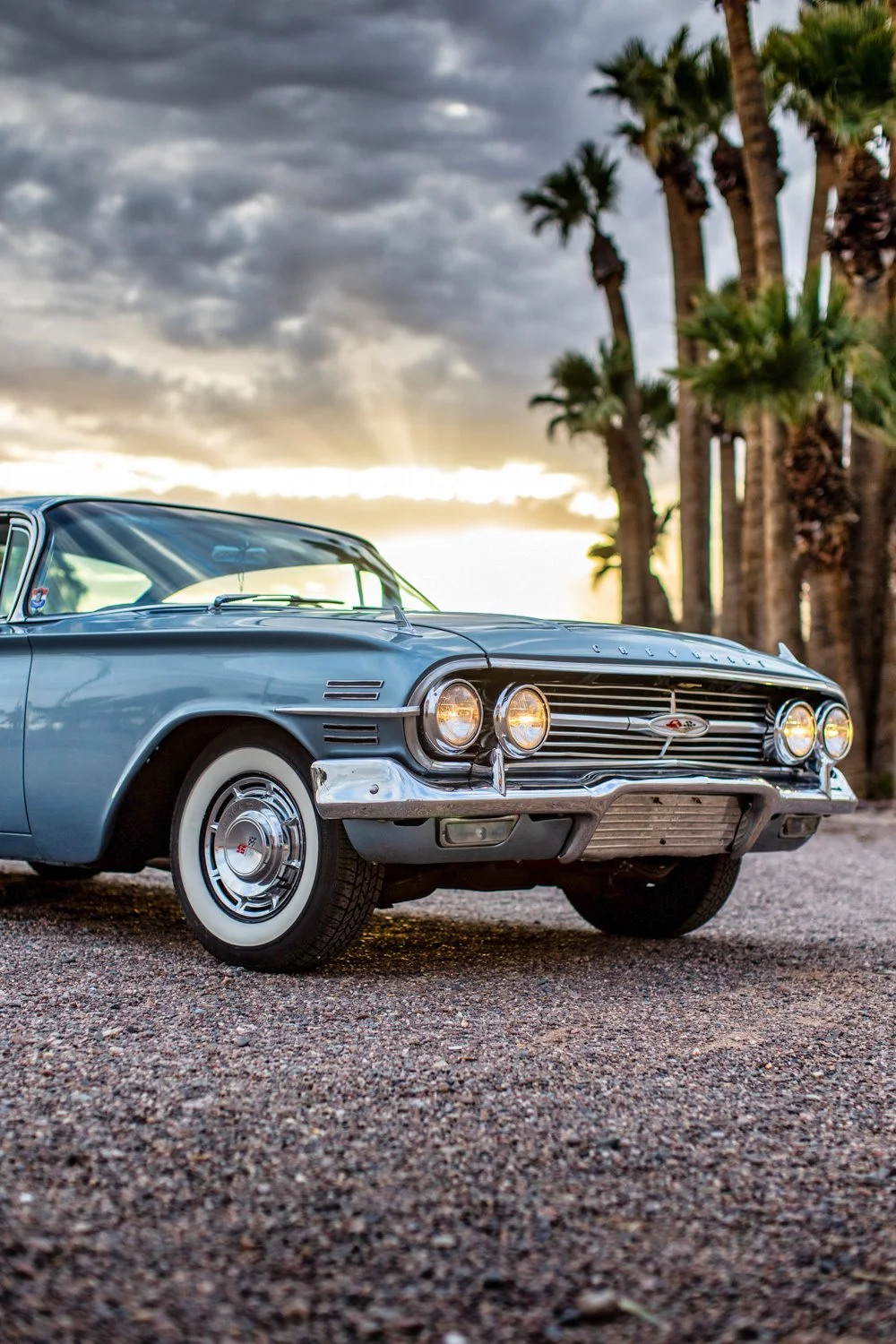 A vintage blue Chevrolet car parked on a gravel surface with palm trees in the background at sunset.