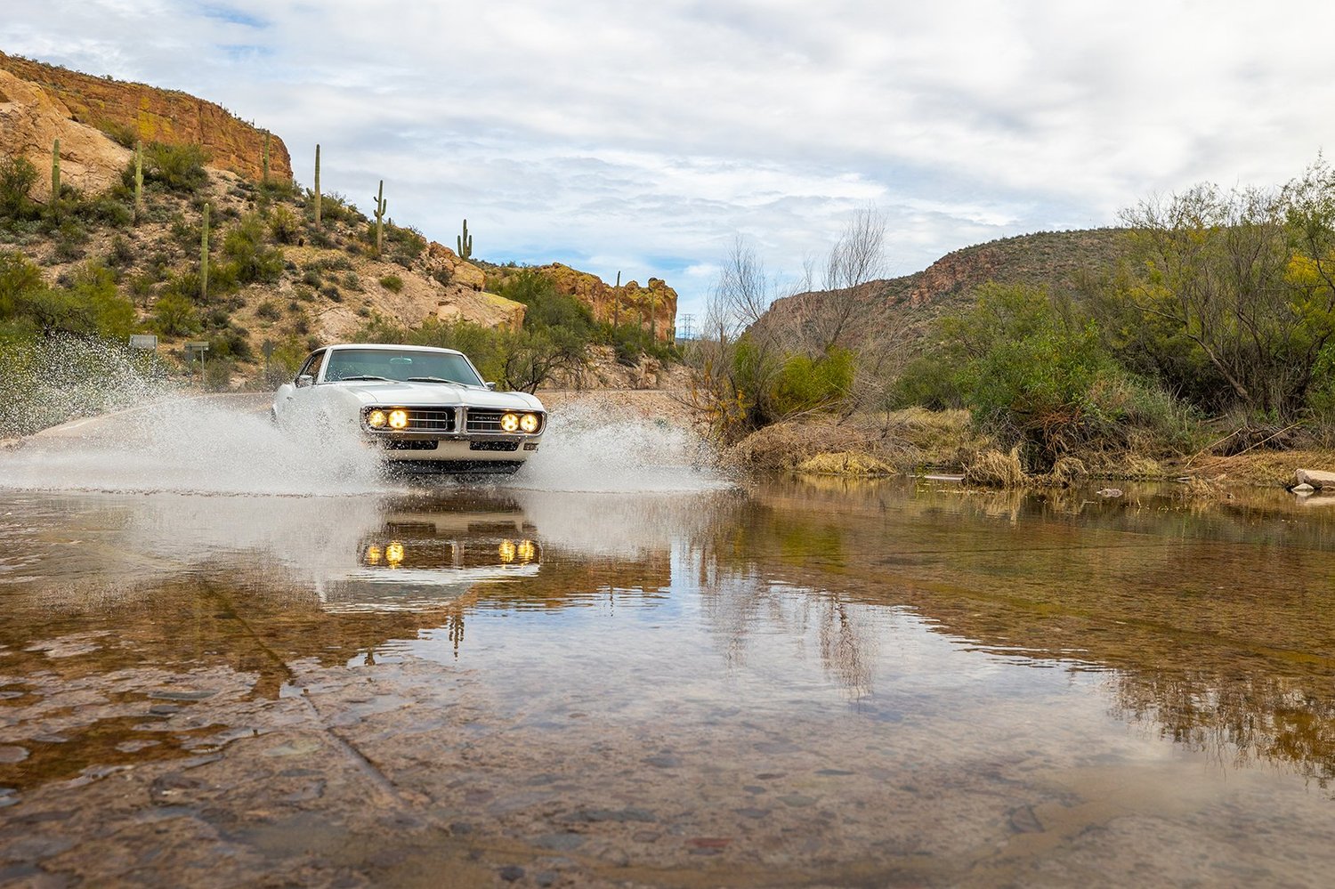 A classic silver car driving through a shallow creek in a desert landscape with cacti and rocky hills under a cloudy sky.
