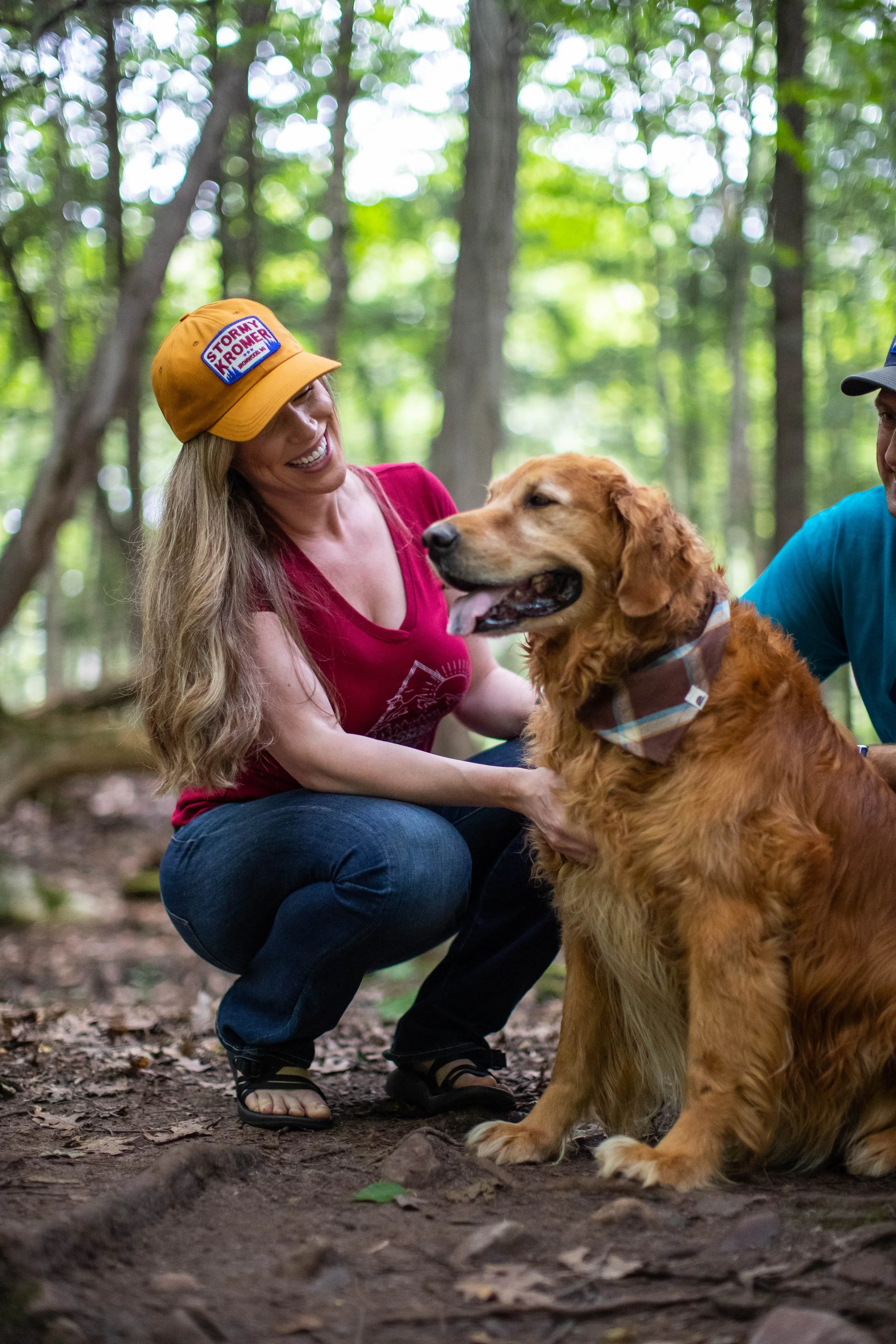 A woman with long blonde hair wearing a yellow hat and red shirt smiling while petting a large golden retriever in a wooded area.