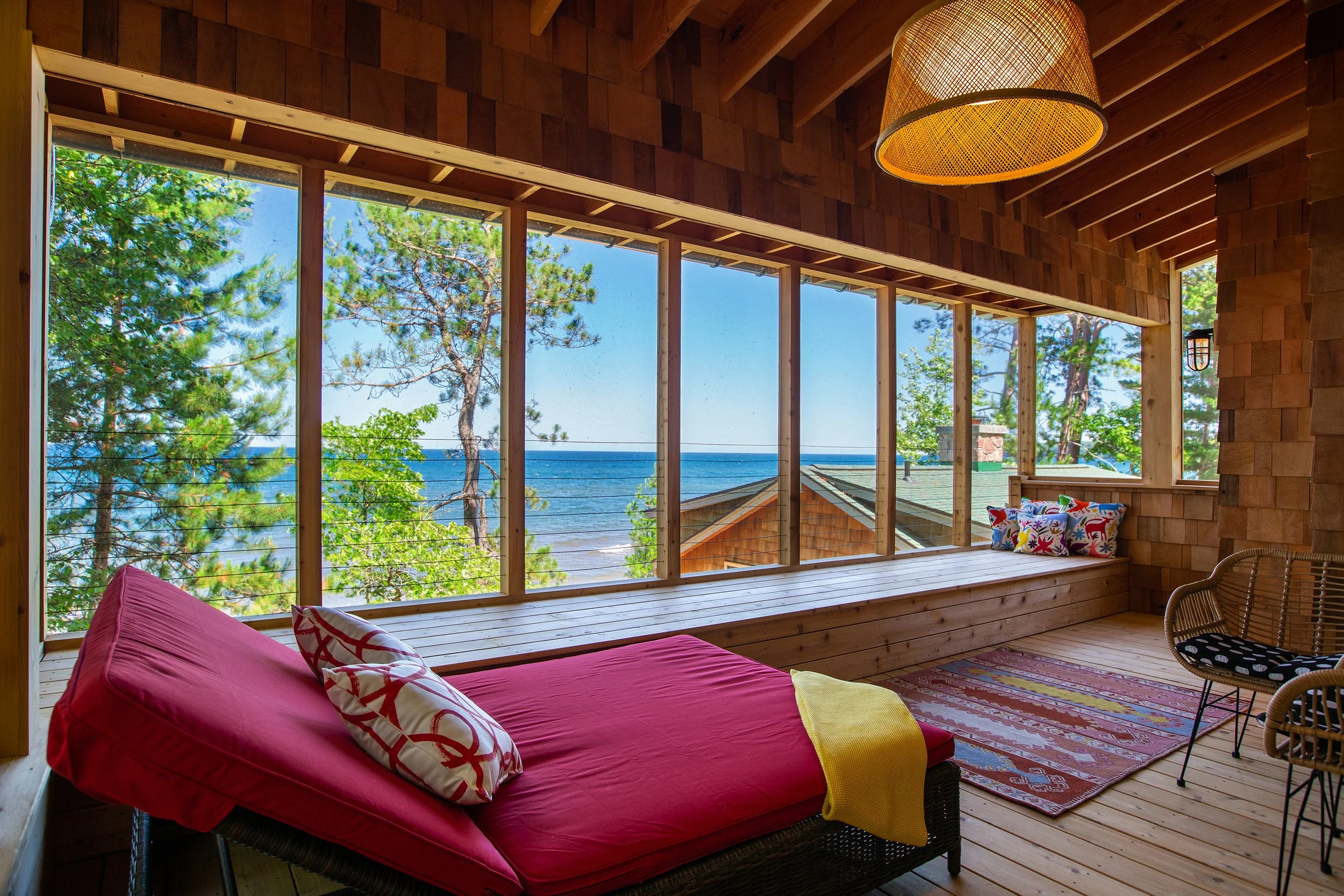 A cozy indoor space with a view of the ocean through large windows, featuring a red daybed with decorative pillows, a woven hanging light, colorful rug, and wicker chairs, creating a relaxed seaside atmosphere.
