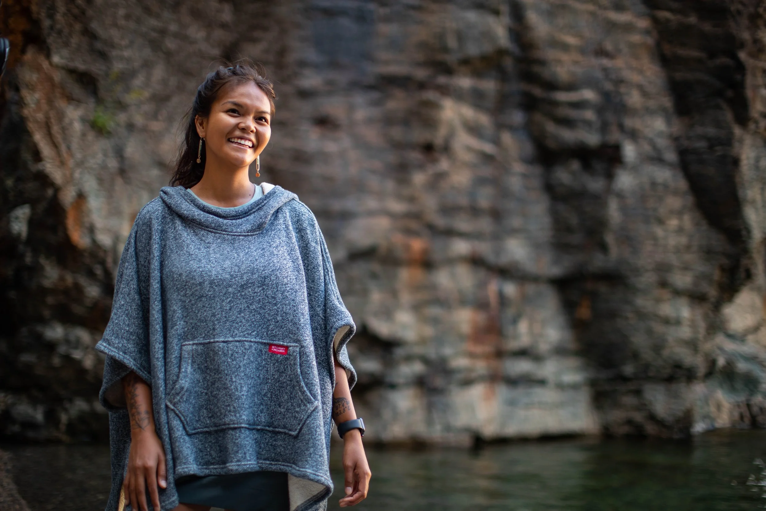 A woman smiling outdoors in front of rocky cliffs, wearing a gray poncho and earrings.