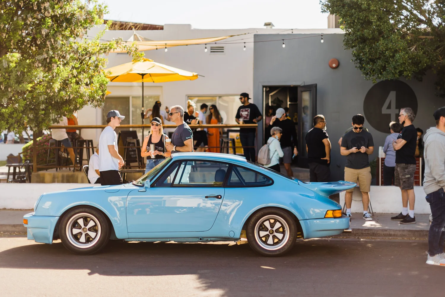 A vintage blue sports car parked on the street outside a cafe or restaurant with people socializing, some looking at their phones, others talking, and a few in line at the entrance.