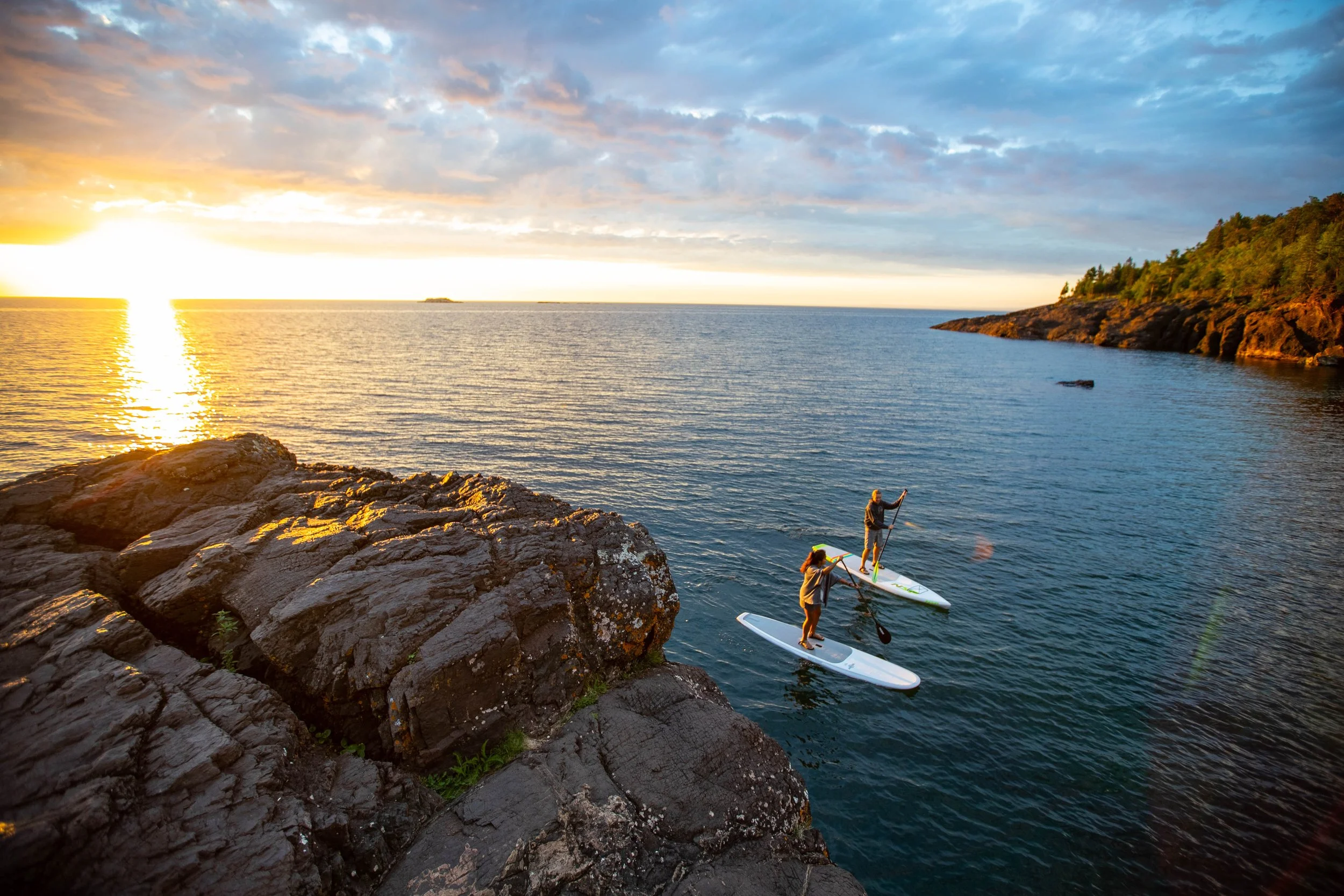 Two people stand on paddleboards near a rocky shoreline during sunset, with the sun low on the horizon and a partly cloudy sky.