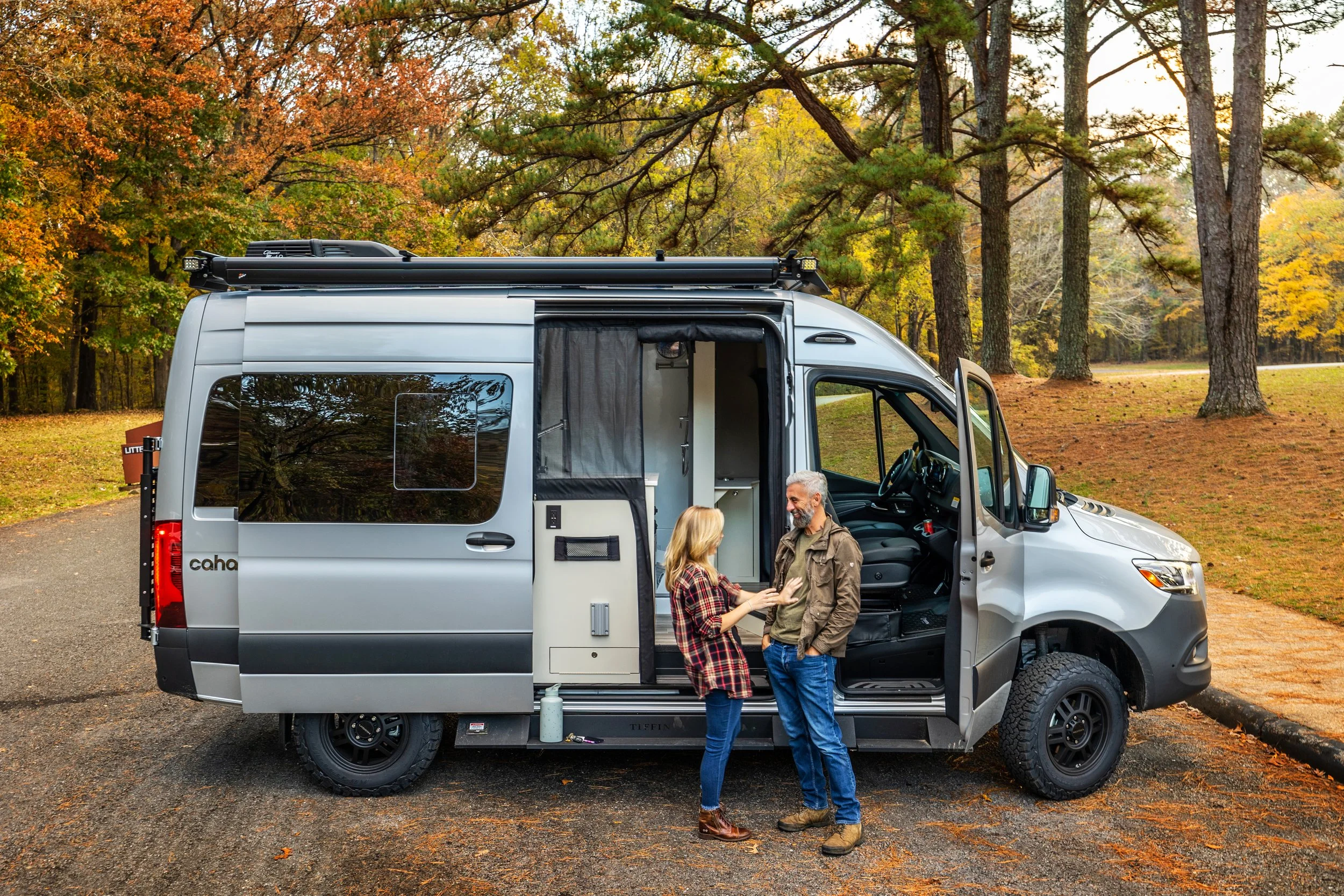 A couple standing outside a silver camper van in a wooded park with fallen leaves, talking and smiling.