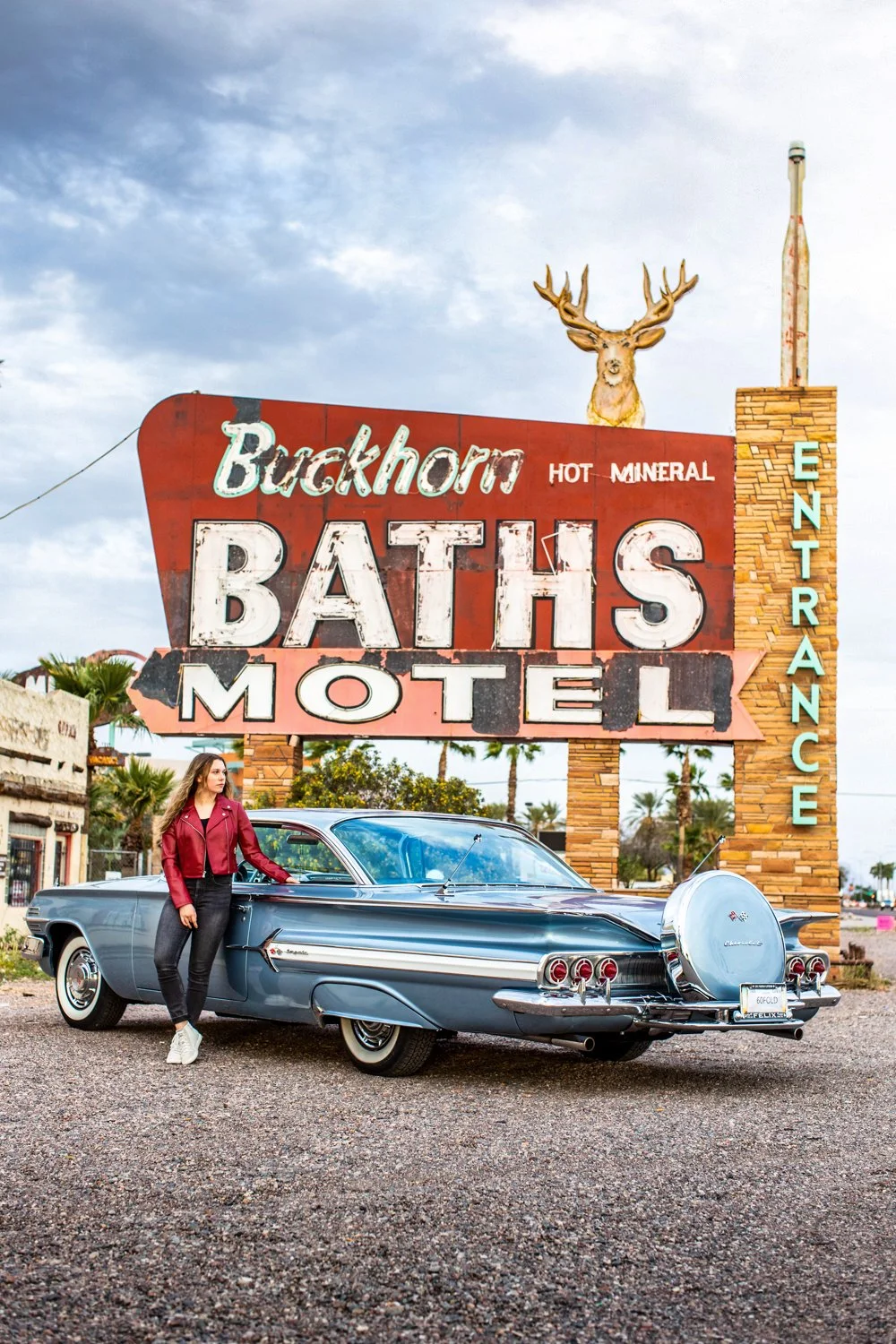 A woman in a red jacket and black jeans leaning on a vintage blue car parked in front of a large retro motel sign reading 'Buckhorn Baths' with a mounted deer head on top. The sign advertises hot mineral baths and the motel's entrance in the backgrou
