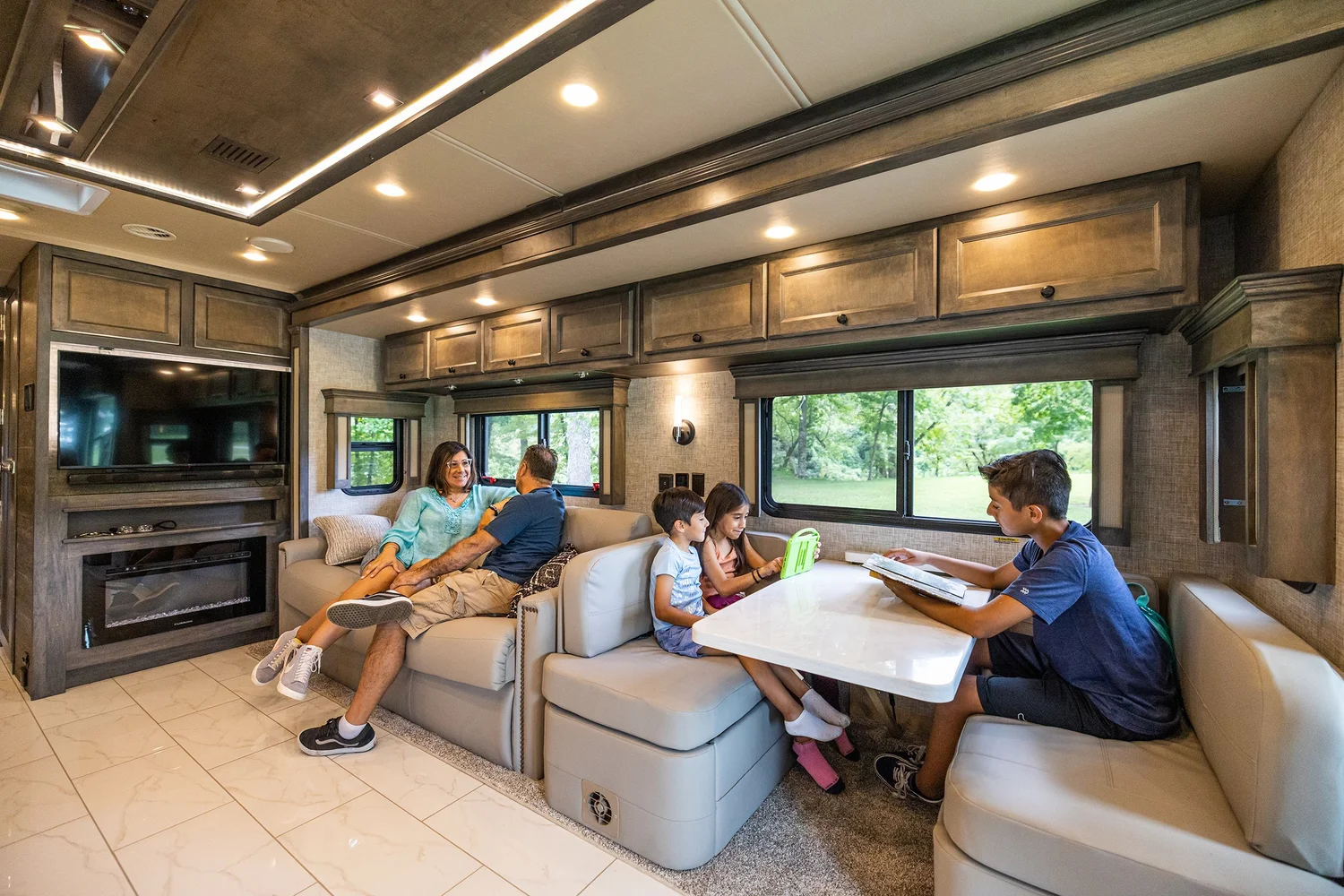 View of the interior of a recreational vehicle with a family relaxing on a sofa and children at a dining table, large windows showing trees outside, and a TV mounted on the wall.