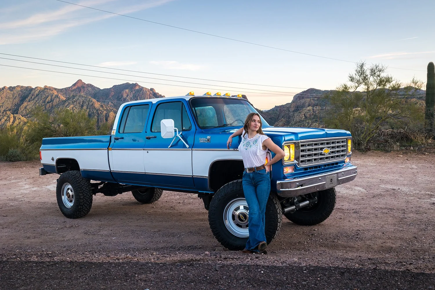 A woman leans against a blue and white vintage Chevrolet pickup truck in a desert landscape with mountains, cactus, and sparse trees in the background during sunset.