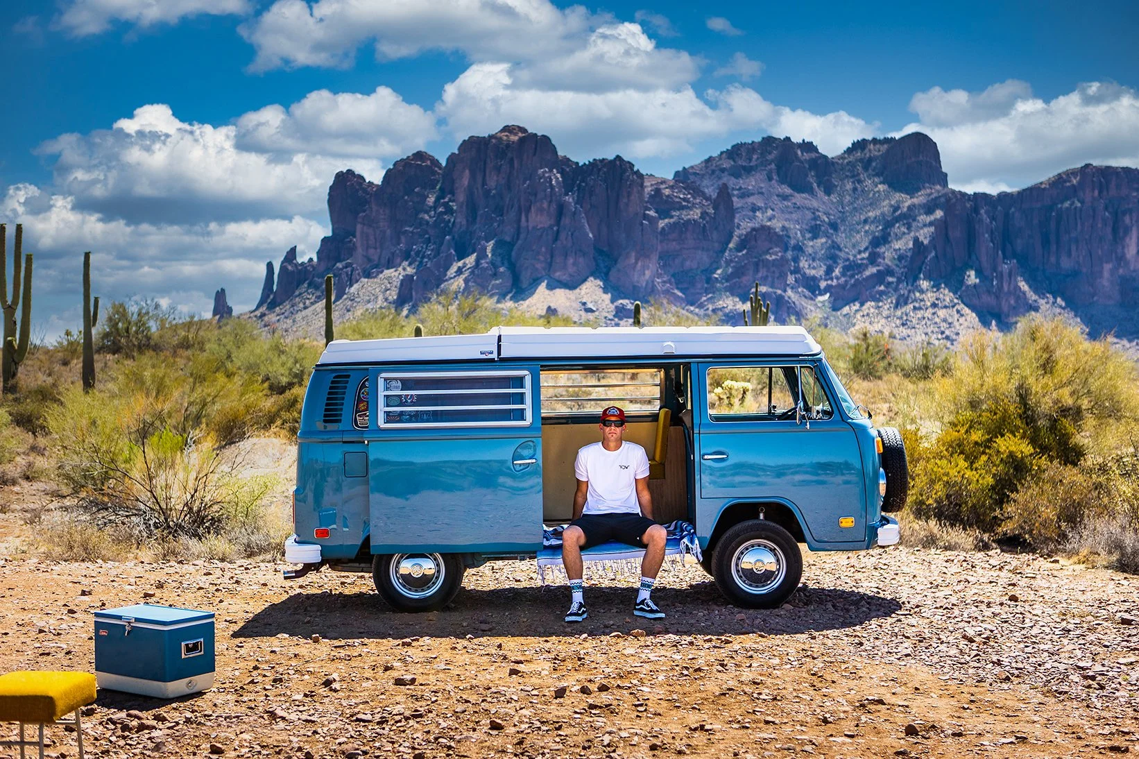 A man sitting on the step of a blue vintage van parked in a desert landscape with cacti and mountains in the background.