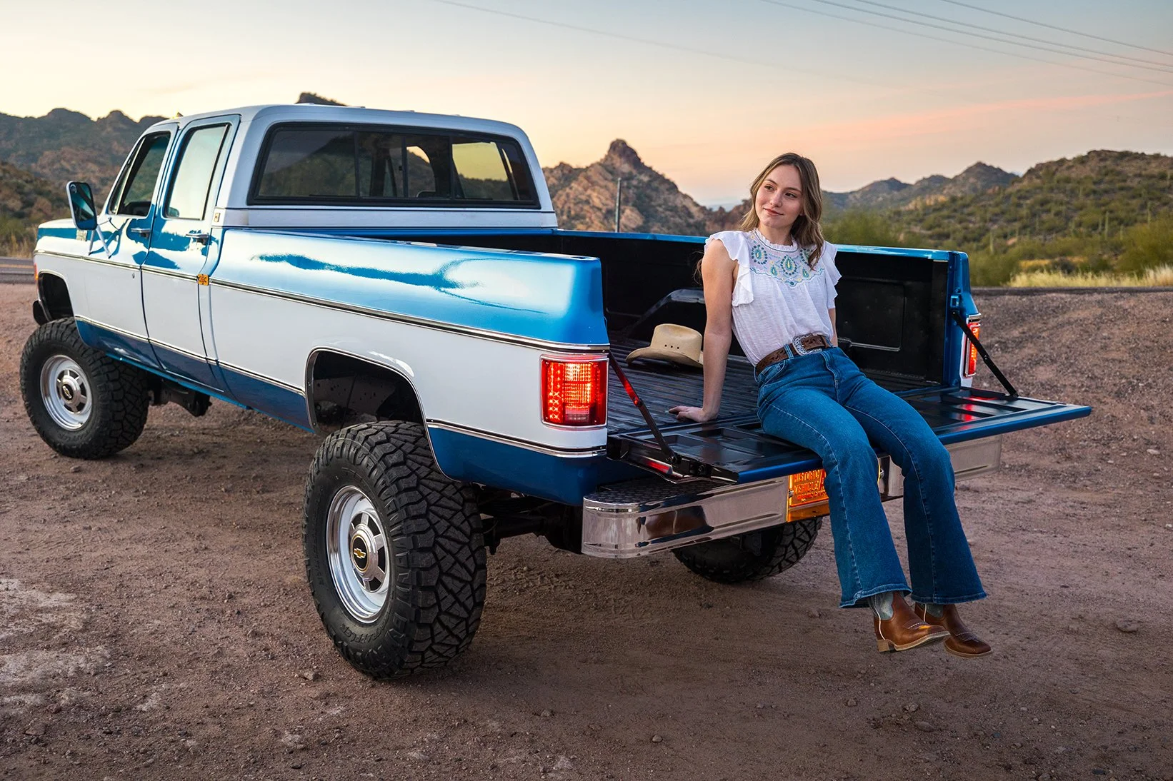 A woman sitting on the open tailgate of a blue and white pickup truck in a desert landscape during sunset, with mountains in the background, wearing a white blouse, blue jeans, and brown boots.