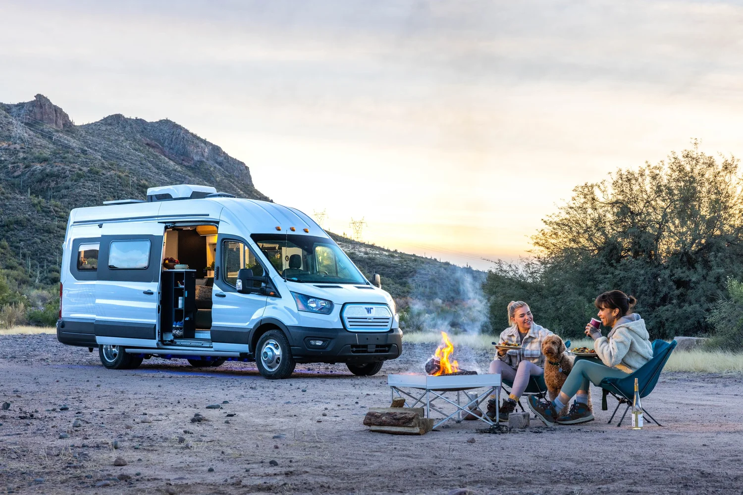 Two women sitting by a campfire with a small dog, enjoying food and drinks next to a white camper van in a desert landscape at sunset.