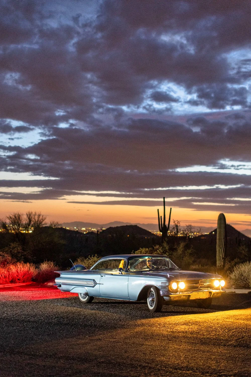 A vintage silver car parked on a desert road during sunset, with saguaro cacti in the background and dramatic clouds in the sky.