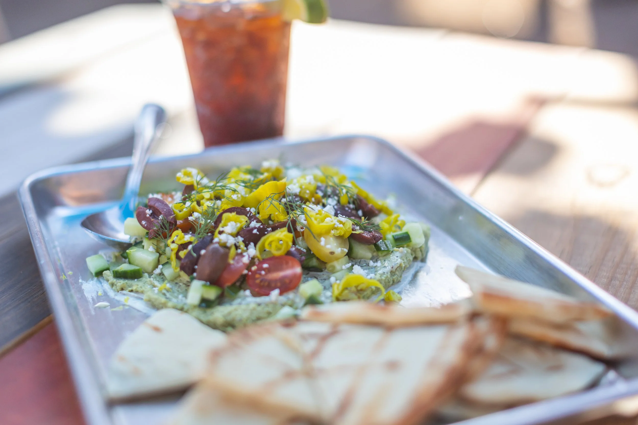 A metal tray with a vegetable pizza topped with cherry tomatoes, cucumbers, olives, and yellow peppers, along with a dollop of white sauce and atop a wooden table. In the background, there is a glass of iced tea with lime.