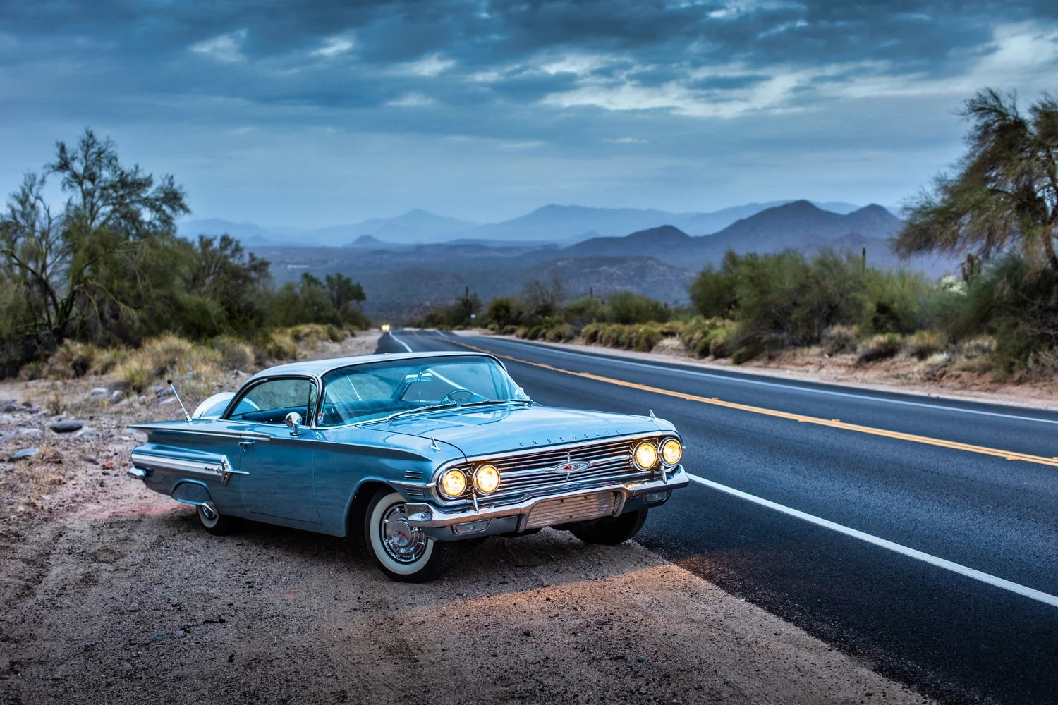 A vintage blue car parked on the side of a deserted road with mountains and cloudy sky in the background.