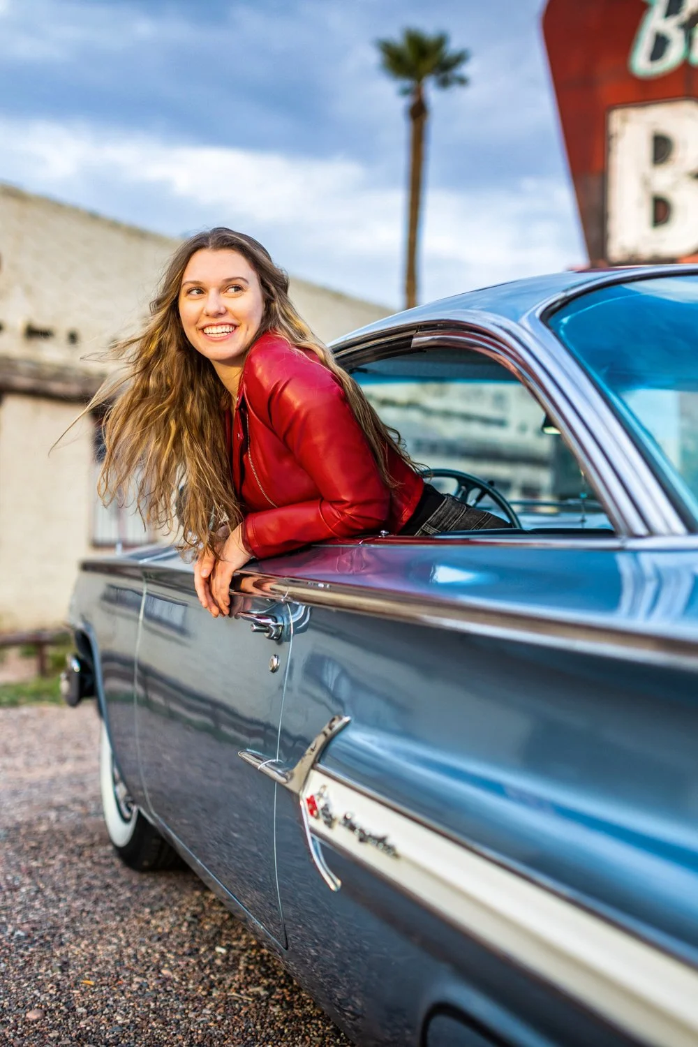 A young woman in a red leather jacket leaning out of a vintage blue car with a happy expression, with a palm tree and a cloudy sky in the background.