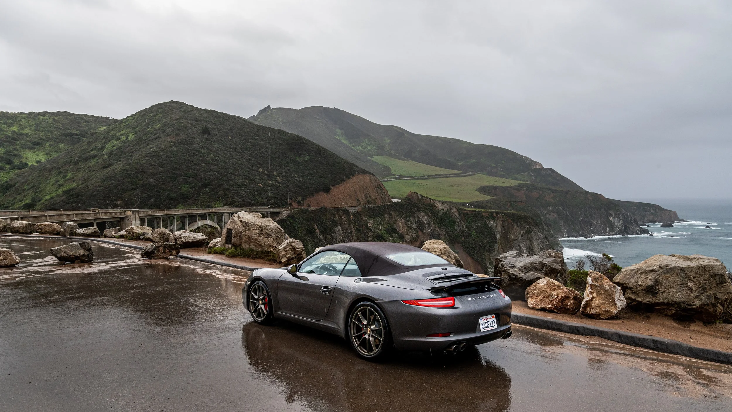 A gray Porsche convertible parked on a wet coastal overlook with rocky shoreline, green hills, and cloudy sky in the background.