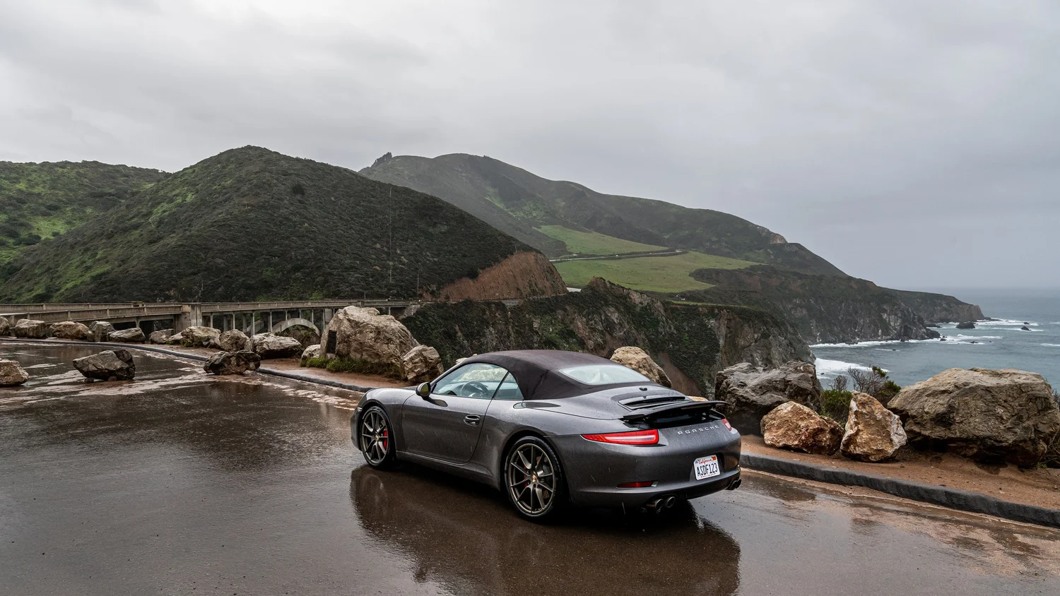 A gray Porsche convertible parked on a wet coastal overlook with rocky shoreline, green hills, and cloudy sky in the background.