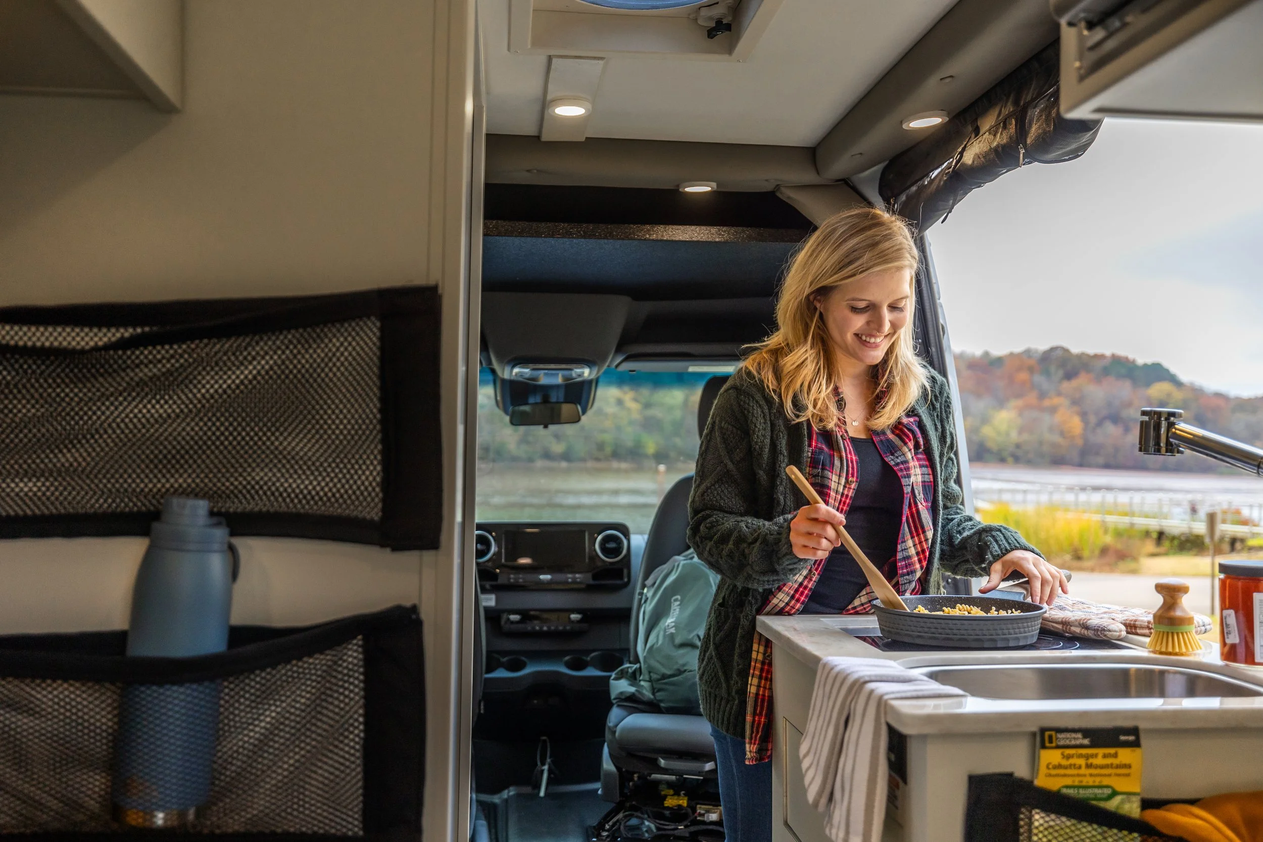 A woman cooking inside a camper van with a scenic view of a lake and trees with fall foliage outside the window.