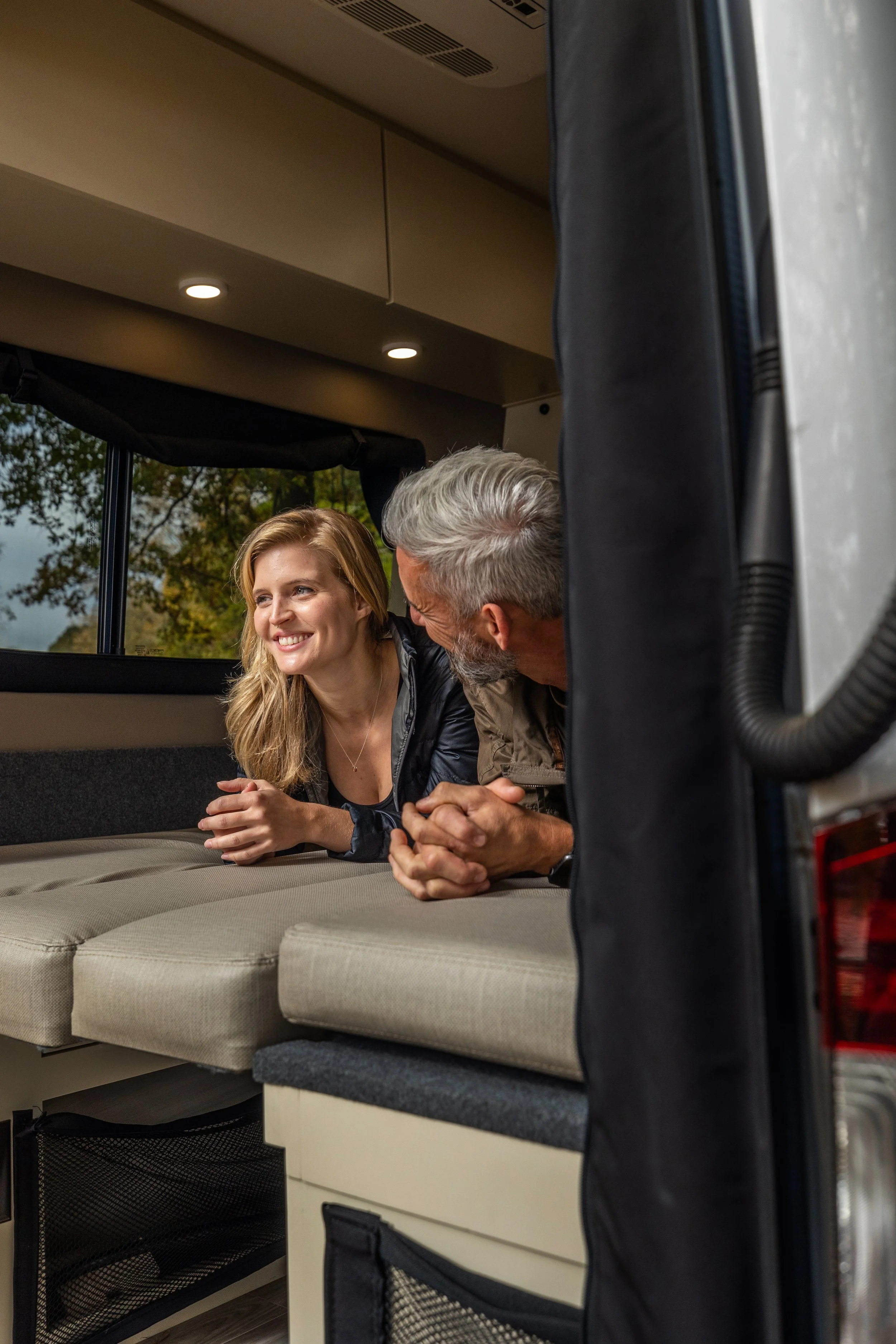 A young woman and an older man lying on their stomachs inside a camper van, smiling and talking to each other, with the outside scenery visible through the window.