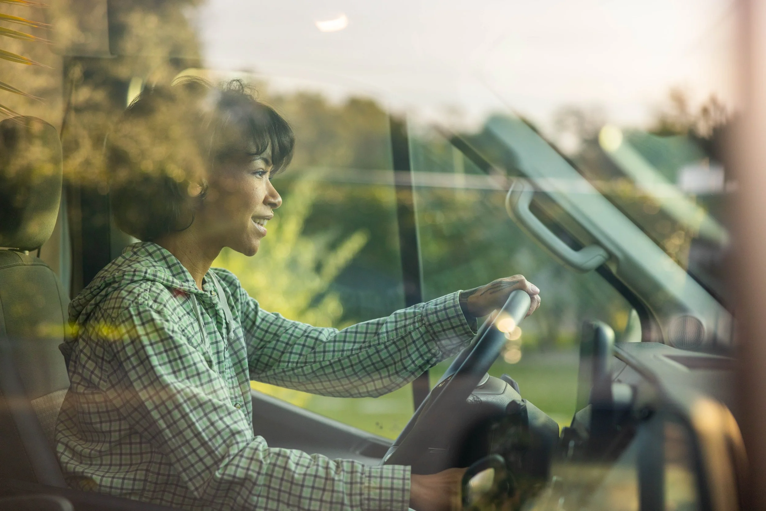 Woman driving a vehicle, viewed through the front windshield with greenery outside.