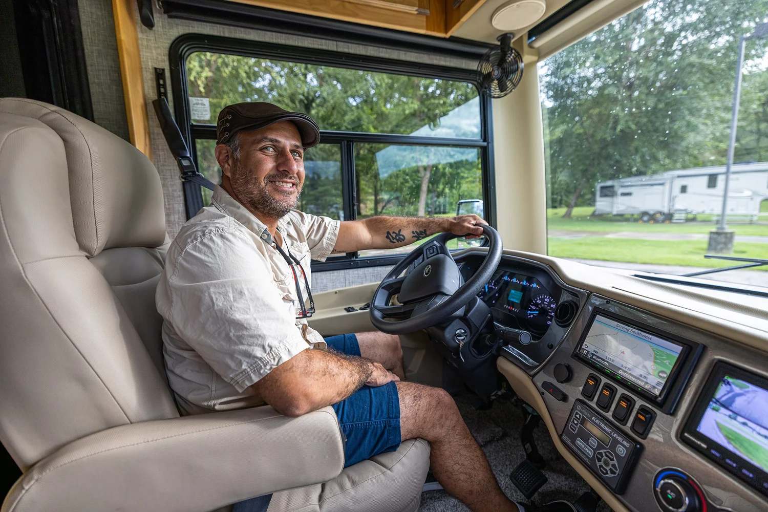 A man sitting in the driver's seat of a recreational vehicle (RV), smiling, with a scenic view of trees and other RVs outside the window.